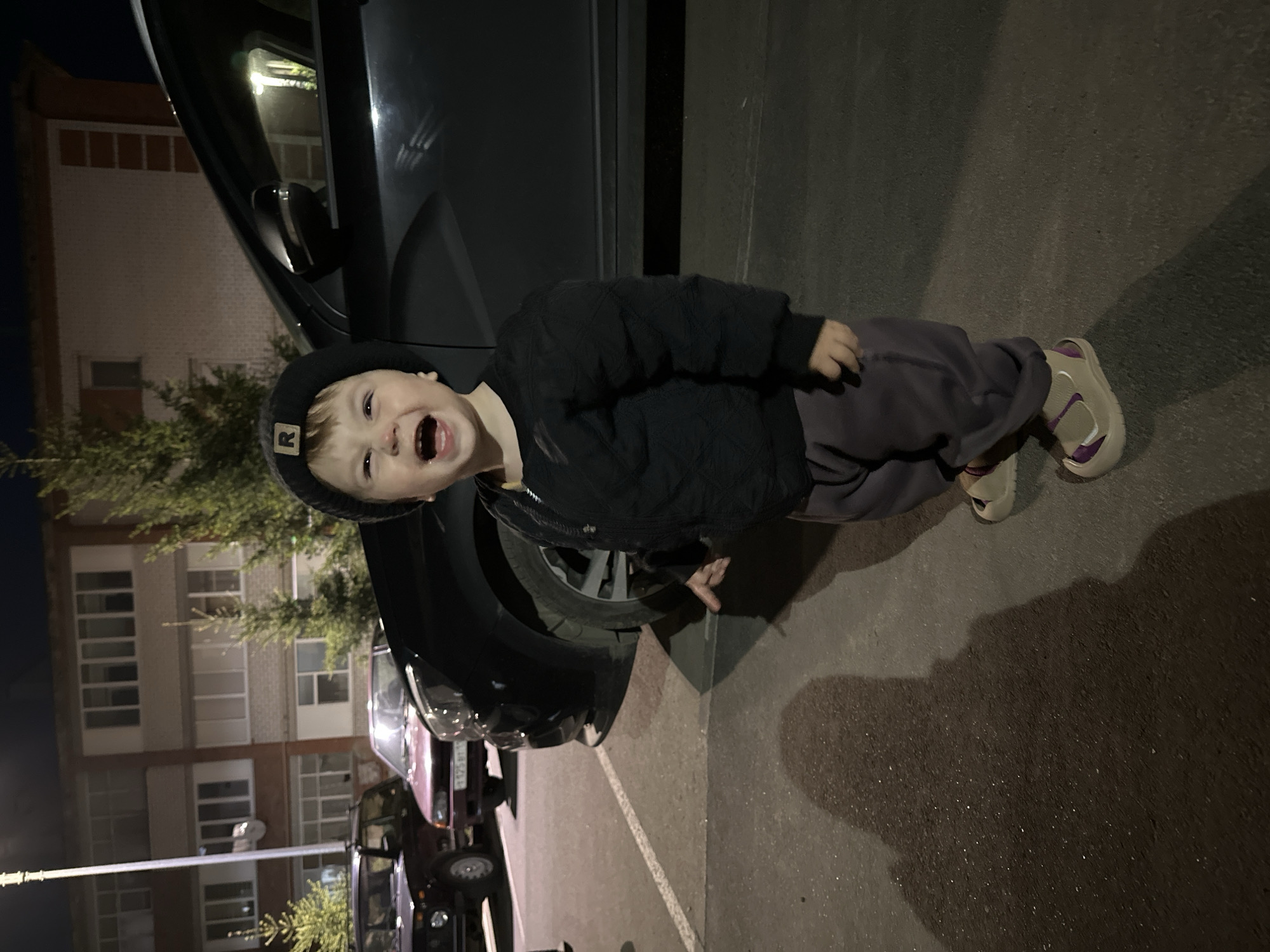 A young child, likely a toddler, is standing in a parking lot or paved area next to a dark-colored car. The child is wearing a dark hat with a logo, a dark quilted jacket, and dark pants, along with light-colored sandals with purple accents. The child is facing the camera with an open-mouthed expression. To the left, the corner of an apartment building is visible with multiple rows of windows. Streetlights are present. Other vehicles are parked in the background. The location is Высокая Гора, Russia.