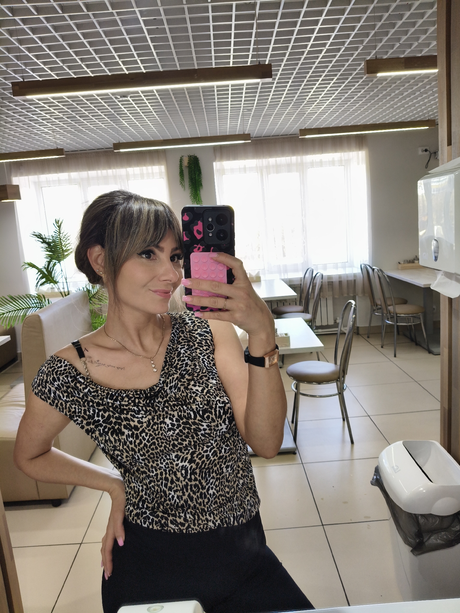 A woman is taking a selfie in what appears to be a brightly lit office or cafeteria space. She is holding a pink and black phone up to her face. She has brown hair, bangs, and is wearing a leopard print top and black pants. There are several tables and chairs in the background, along with a large window providing natural light. A tall plant stands near a sofa in the background. The walls are light-colored, and the flooring is tiled. The location is in Chaikovsky, Russia.