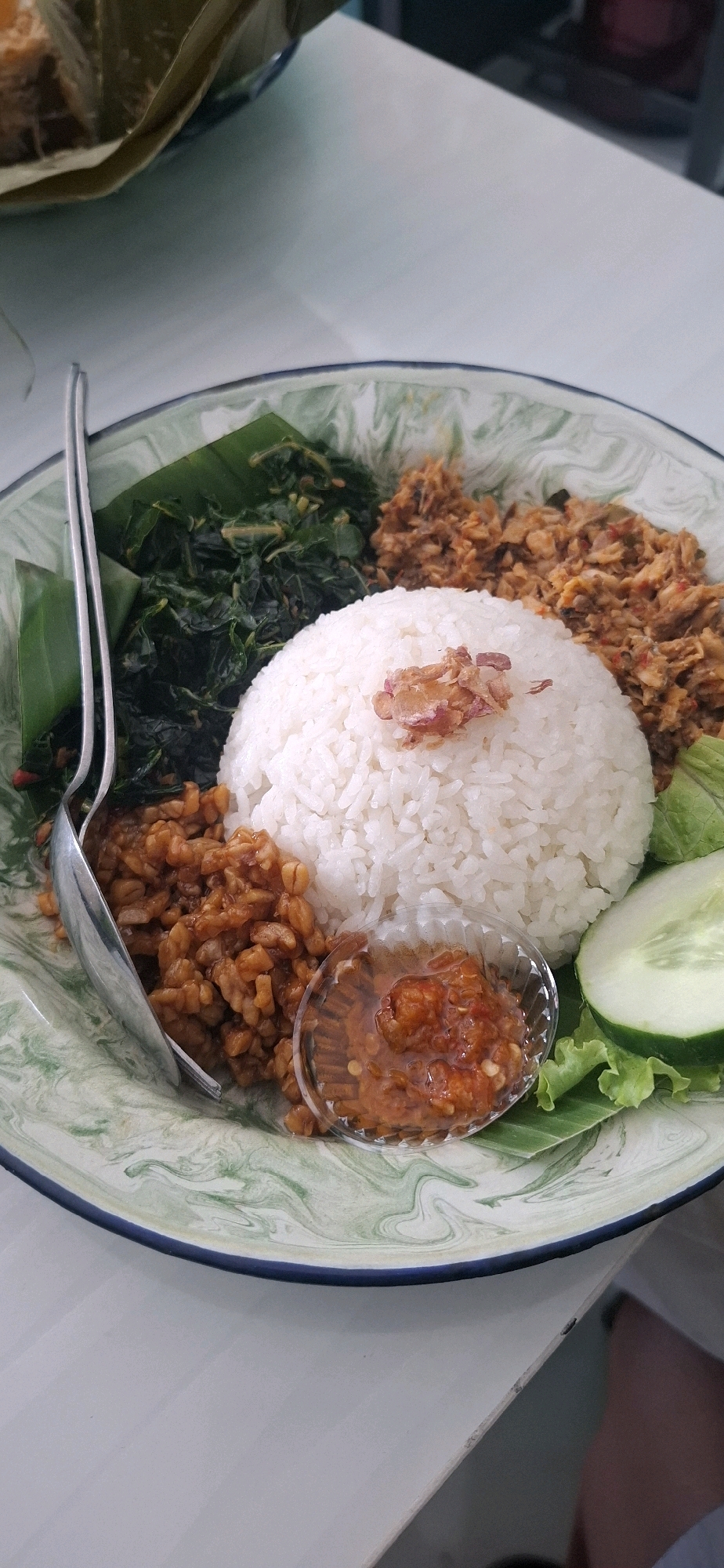 A close-up, overhead shot shows a plate of Nasi Uduk, a fragrant rice dish, served with various accompaniments on a light-colored, marbled plate. The dish is centered, with a large mound of white rice taking prominence. To the left of the rice is a pile of dark green cooked leafy vegetables, possibly spinach or kale, arranged on a banana leaf. Next to the vegetables and to the left of the rice is a serving of tempeh, a fermented soybean cake, cut into small cubes and stir-fried with a savory sauce. A shiny metal spoon rests on the left side of the plate, partly covering the tempeh.
To the right of the rice, there are two portions of what appear to be shredded or flaked meat, likely fish or chicken, seasoned with a reddish spice blend. Below these meat portions, a slice of cucumber and some lettuce leaves are placed as garnishes. In the foreground, nestled between the rice and tempeh, is a small, clear plastic container filled with a vibrant orange sambal, a spicy chili paste. The background is blurred, but suggests an indoor setting with a light-colored surface, possibly a table. The overall presentation indicates a served meal, likely in a restaurant or home setting in Surakarta, Indonesia.