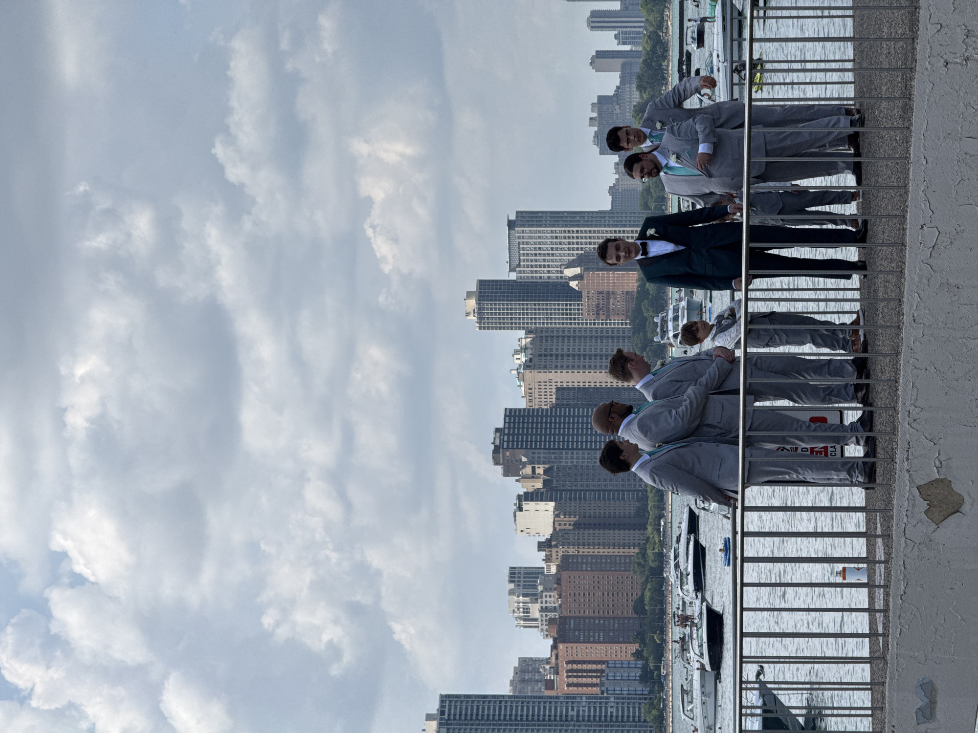 In Chicago, USA, eight individuals, including seven men and one boy, are positioned on a promenade with a metallic railing. The men are formally dressed in suits and tuxedos, mostly in shades of gray, with some wearing teal ties and pocket squares. One man is in a dark green tuxedo with a black bowtie. The boy is also in a gray suit.
Behind them, a body of water, likely Lake Michigan, is visible, dotted with several white motorboats and a few distant people. The city skyline of Chicago forms the backdrop, characterized by numerous high-rise buildings of varying architectural styles and colors. The sky above is filled with a mix of bright white and gray clouds, suggesting overcast or partially cloudy weather. The promenade itself is a concrete surface, with visible peeling paint in the foreground.