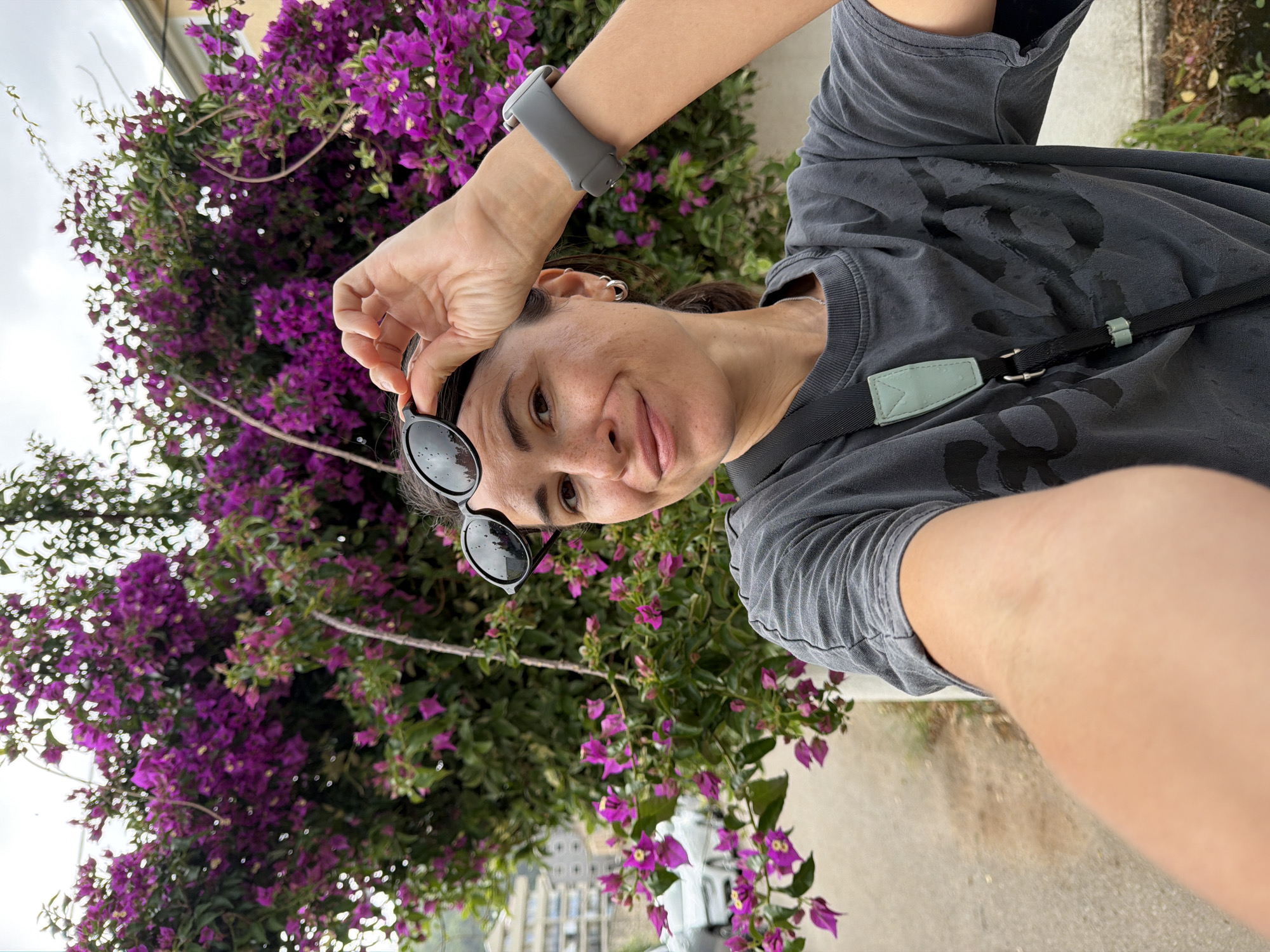 A woman takes a selfie, holding dark sunglasses on top of her head with her right hand, while looking directly at the camera with a subtle smile. She has dark hair pulled back, wears a grey t-shirt with dark, indistinct graphic elements on the front, and a grey smartwatch on her left wrist. Small hoop earrings are visible in her left ear. A black shoulder strap with a light green rectangular accent crosses her chest.
The background is dominated by lush, vibrant purple bougainvillea flowers and green foliage, suggesting a warm, possibly Mediterranean, environment. Below the floral canopy, on the left side, are glimpses of white buildings and rooftops, indicating a town or city. The sky is bright and overcast, diffusing the light. The lower right foreground shows a light-colored paved surface. The image captures a casual, relaxed moment during the daytime.