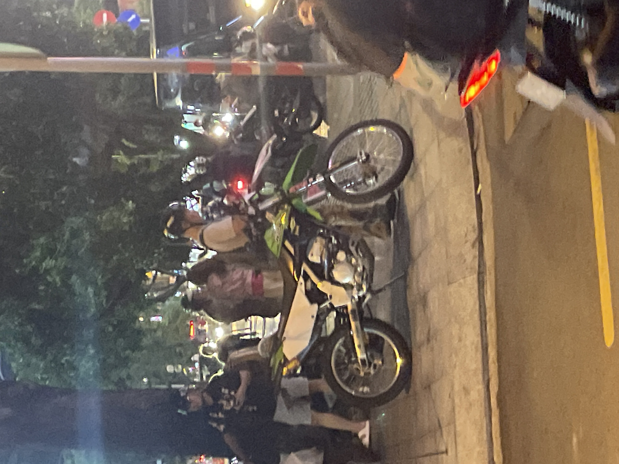 A street scene at night in Thủ Đức, Vietnam, features a green and white dirt bike parked on a sidewalk. The bike is the primary focus, with its front wheel turned slightly to the left. In the background, several people are visible, some standing and others on motorbikes. Trees and streetlights create a dimly lit atmosphere. A traffic light with red and blue signals is visible on a pole to the right of the frame. Other vehicles, including a dark-colored van or SUV, are present further down the street. The ground is paved with concrete and asphalt, and there is a yellow line painted on the road. The image appears to be taken from a slightly elevated perspective.