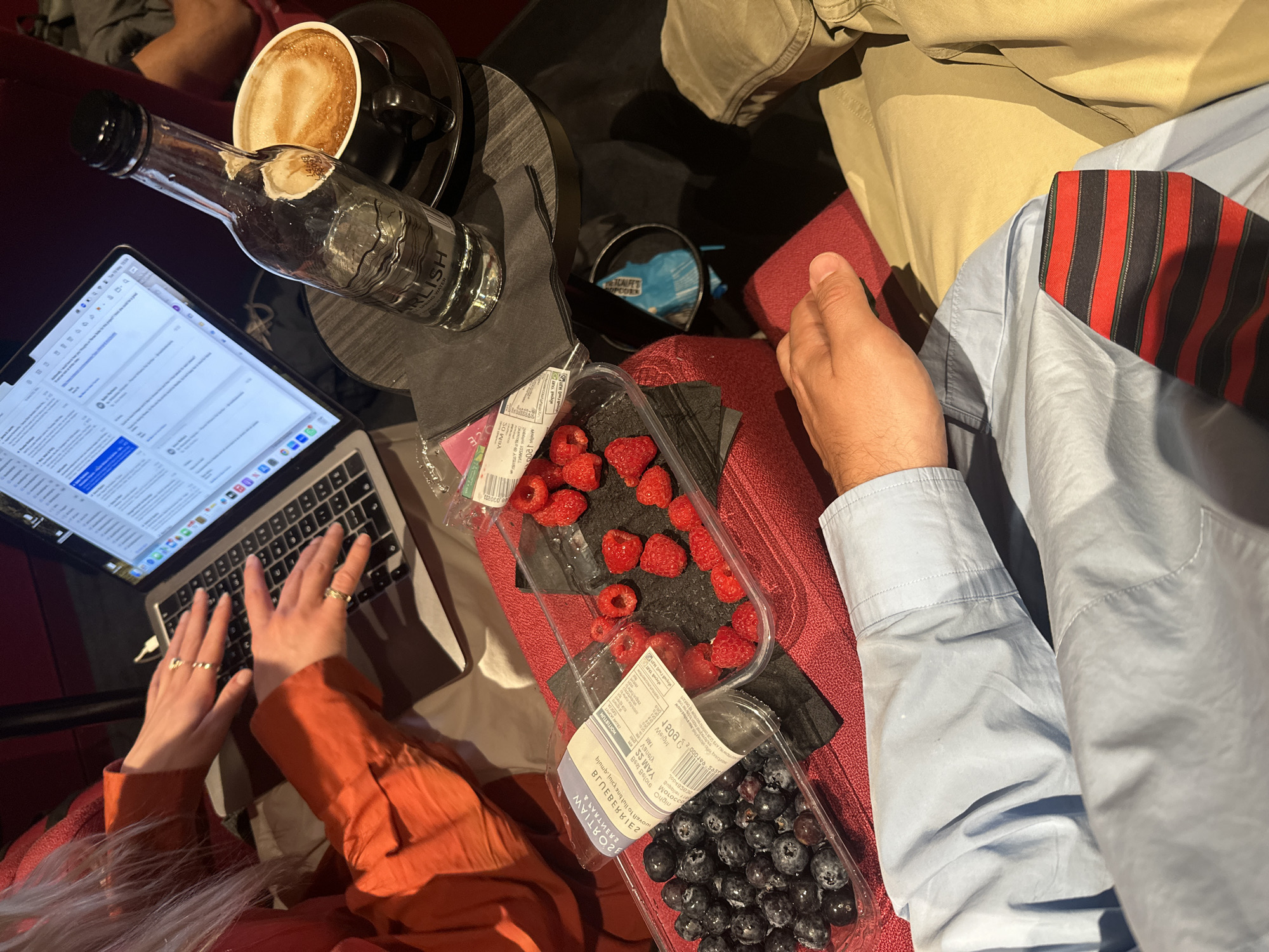 A top-down indoor shot depicts two individuals seated in adjacent chairs. The person on the left is working on a laptop, their hands poised on the keyboard. They are wearing an orange-colored top and have blond hair. To their right are containers of raspberries and blueberries on a black napkin. A glass bottle of water, labeled "Carlisle Spring Water," and a cup of coffee sit on a black side table near the laptop.
The person on the right is wearing khaki pants and a light blue shirt with a red and black striped tie.
A bag of "Metcalfe's Popcorn" is on the seat between the two individuals. The location is likely a public setting, possibly a theater or lounge, in London, United Kingdom.