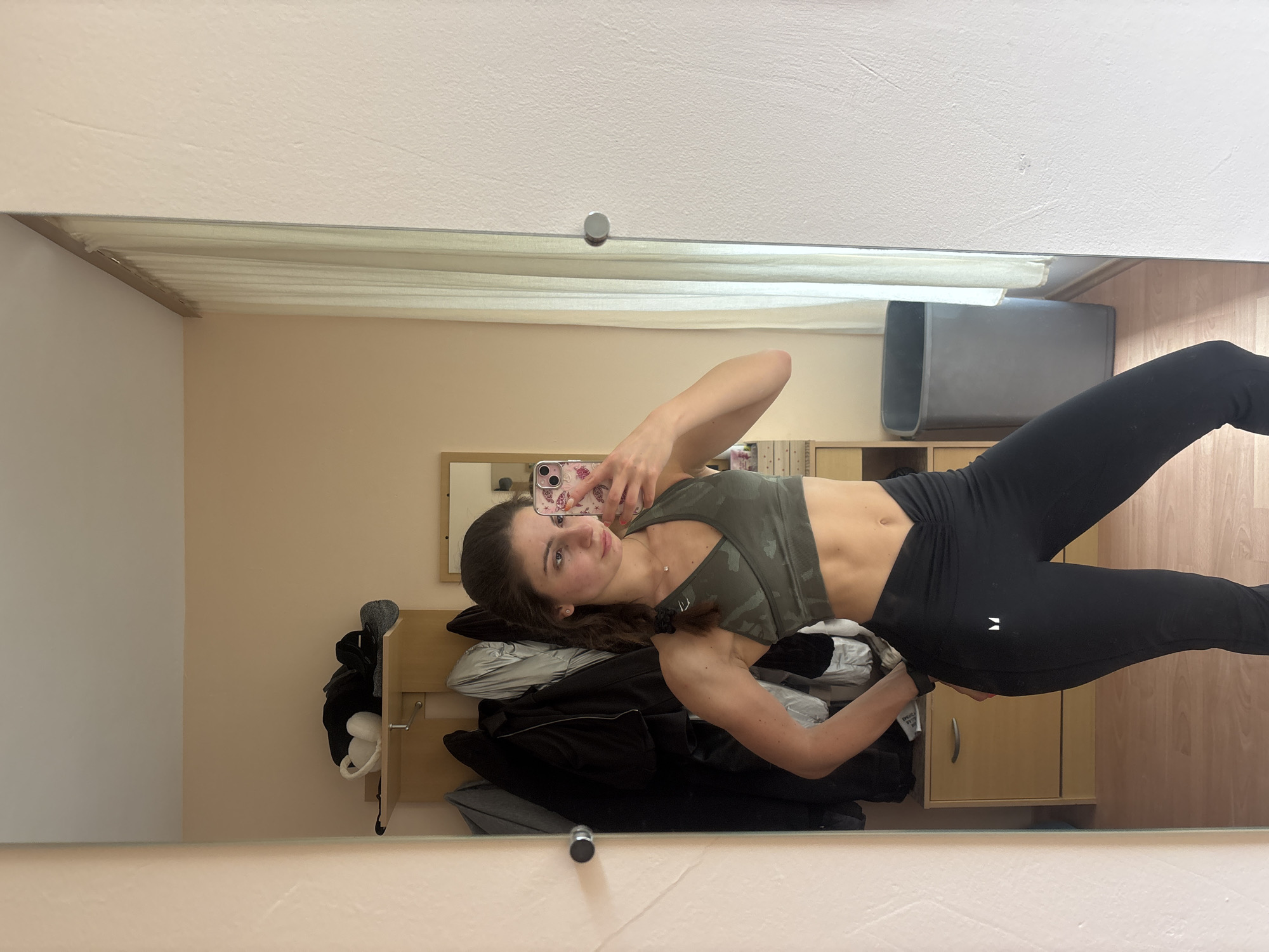 A woman is taking a selfie in a mirror. She is wearing a camo sports bra and black leggings. The mirror reflects a bedroom or dressing area with a coat rack, a storage unit, and a beige wall. The location is likely indoors, potentially in a residential setting, but the city of Varna, Bulgaria, cannot be confirmed from the image. There is no direct evidence in the image to indicate a specific location in Varna or even Bulgaria.