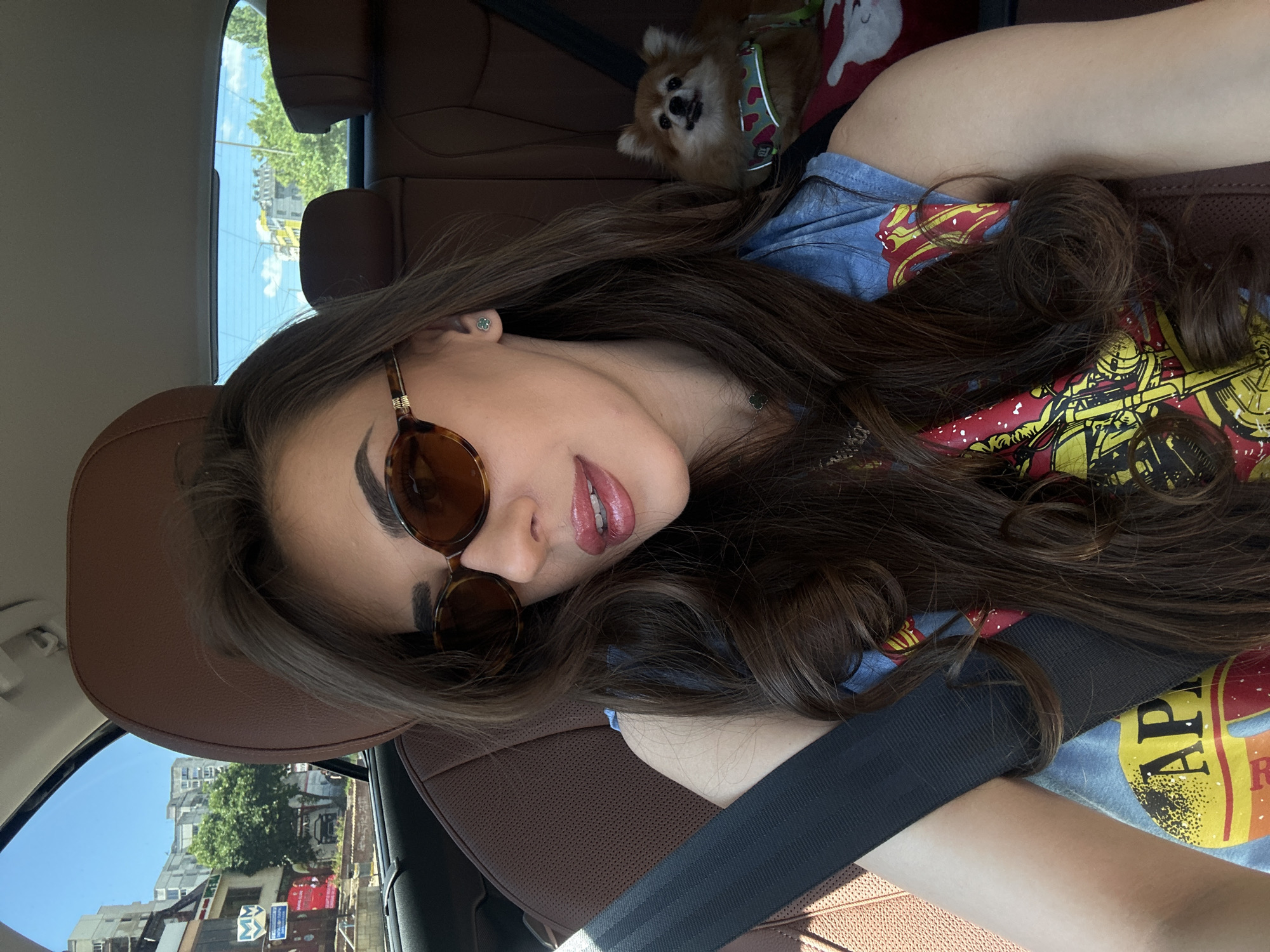 A woman with long, dark brown hair is taking a selfie in the passenger seat of a car. She is wearing sunglasses, a blue t-shirt with a graphic print, and a necklace. Her lips are glossy and slightly parted. Next to her, a small, fluffy tan dog is sitting on the back seat, wearing a red and green harness decorated with small hearts. The car interior is brown leather. Outside the car window, buildings, trees, and a blue sky with some clouds are visible, suggesting an urban environment in Sofia, Bulgaria.