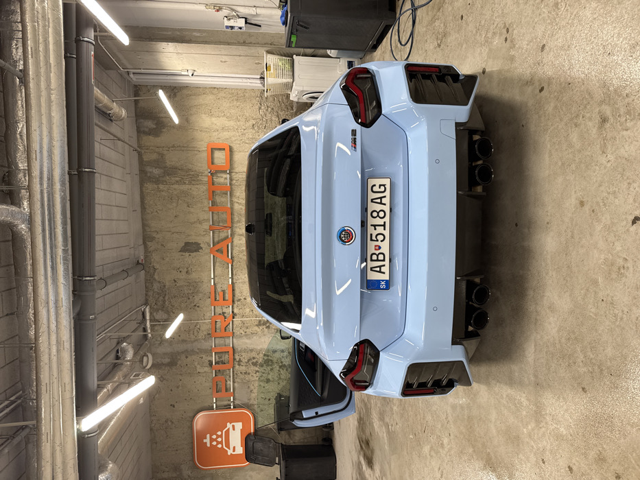 The image displays the rear view of a light blue BMW M2 coupe positioned inside an industrial car detailing or wash facility named "PURE AUTO." The vehicle features a black roof, a custom BMW emblem on the trunk lid, and a Slovakian license plate reading "SK AB-518AG." The driver's side door is open, revealing a light blue trim element within the interior.
The facility's walls are constructed from a textured, worn concrete or stucco, displaying some discoloration. Prominently centered on the back wall is the business name "PURE AUTO" rendered in large, orange, backlit letters. To the left of the name is an orange square sign depicting a car being washed, serving as a logo. The ceiling is exposed, featuring grey ventilation ducts, various pipes, and multiple active fluorescent tube lights providing overhead illumination. The concrete floor exhibits wear, stains, and some moisture.
To the left of the vehicle, a black plastic waste bin is visible. To the right, a white washing machine or similar appliance is situated, topped with a white plastic laundry basket containing green fabric items. Adjacent to this, a dark grey cabinet or trolley is present, with a coiled blue hose or cable lying on the floor in front of it. An electrical outlet or control unit is mounted on the right wall.
This "PURE AUTO" facility is located at Płatnerska 2, 04-207 Warsaw, Poland.