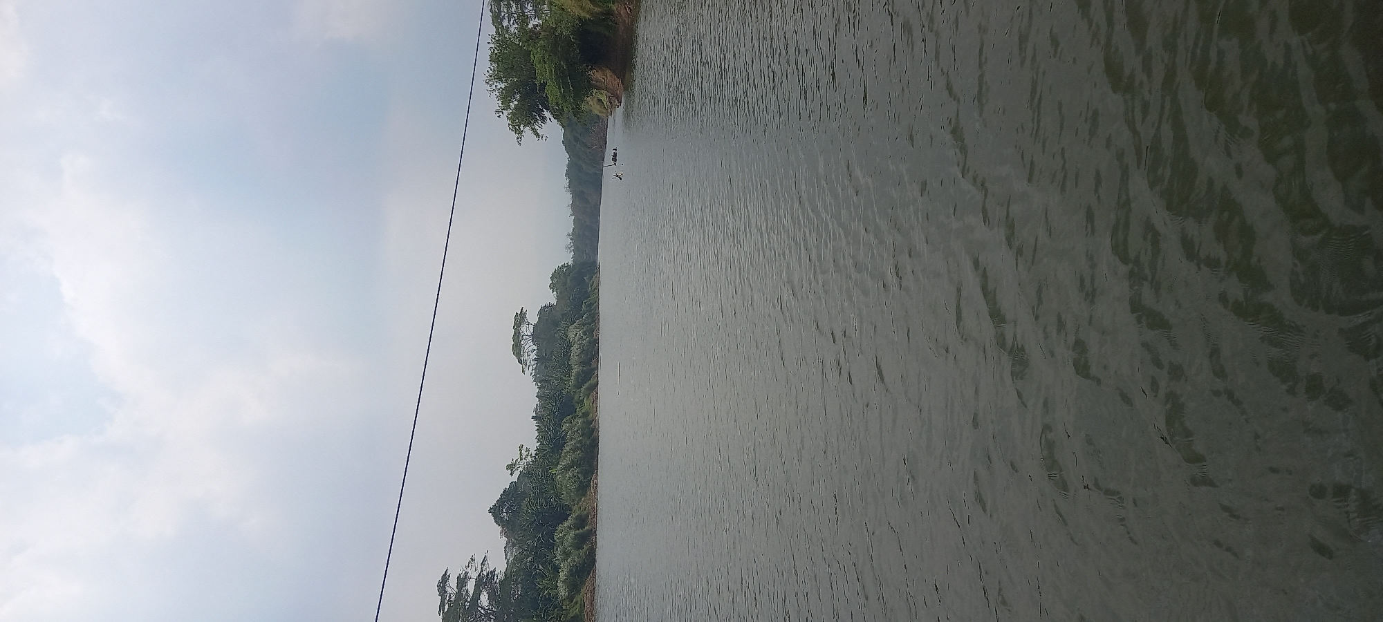 The image shows a wide river with calm water flowing through a natural landscape. On the left bank, lush green vegetation and trees cover the slope, extending into the distance. The right bank is also covered with greenery, with some exposed reddish-brown earth at its base. In the middle of the river, a small, dark structure, possibly a raft or boat with people, is visible. The sky above is overcast with light gray clouds. A thin black wire stretches diagonally across the upper portion of the image, suggesting the presence of overhead power lines. The scene depicts a serene, rural environment, likely near Cintaratu, Indonesia, given the tropical vegetation and the appearance of the river.