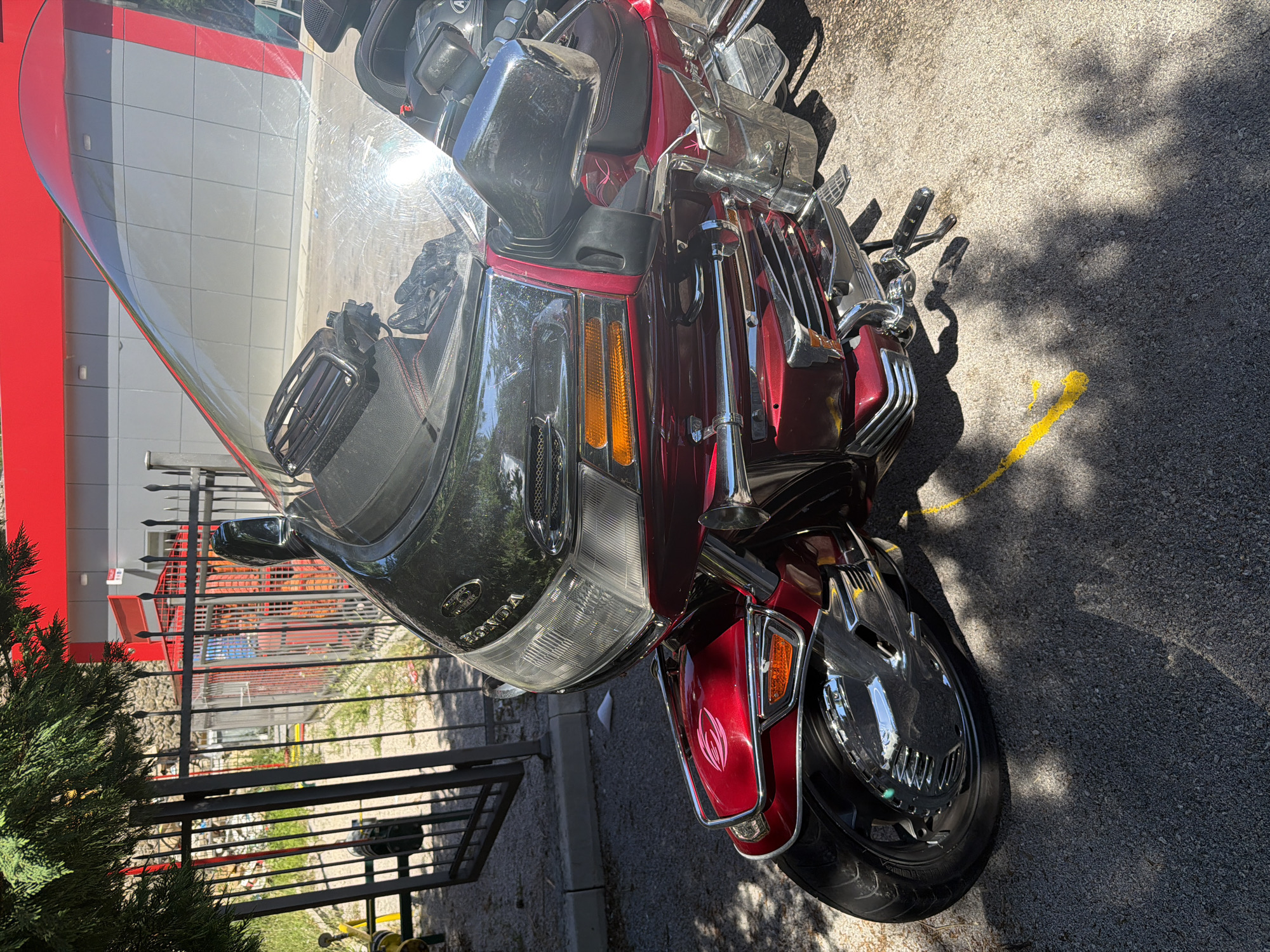 In Sotonići, Montenegro, a red Honda motorcycle is parked on an asphalt surface, partially shaded by trees. The motorcycle is a touring model, characterized by its large fairing, windshield, and ample chrome accents. Its front wheel and part of the lower fairing are visible. The background includes a metal fence, a building with a red and gray facade, and some greenery. A yellow line is painted on the asphalt near the motorcycle.