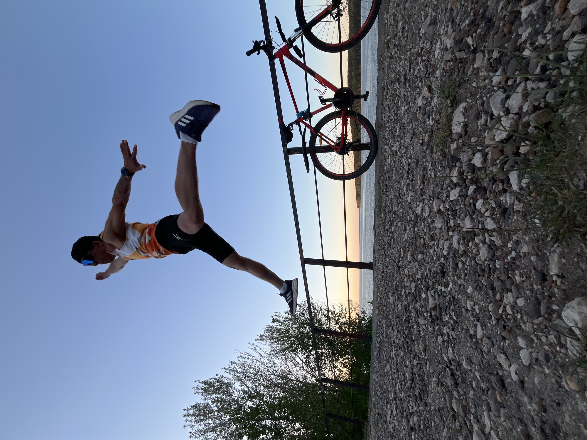 A man is suspended horizontally mid-air, with his right leg extended and right foot connected to a metal railing. His left leg is raised in the air, with a sneaker on his foot. His arms are outstretched, palms facing the viewer. He is wearing sunglasses, a patterned tank top, and black shorts.
Attached to the metal railing is a red mountain bike.
The scene appears to be outdoors, possibly a promenade or observation point. In the background is a body of water and the horizon at sunset. In the foreground is a concrete wall, stones, and vegetation.
The location is assumed to be Chaikovsky, Russia, based on the prompt instructions. The specifics of the exact location within the city are not discernible.