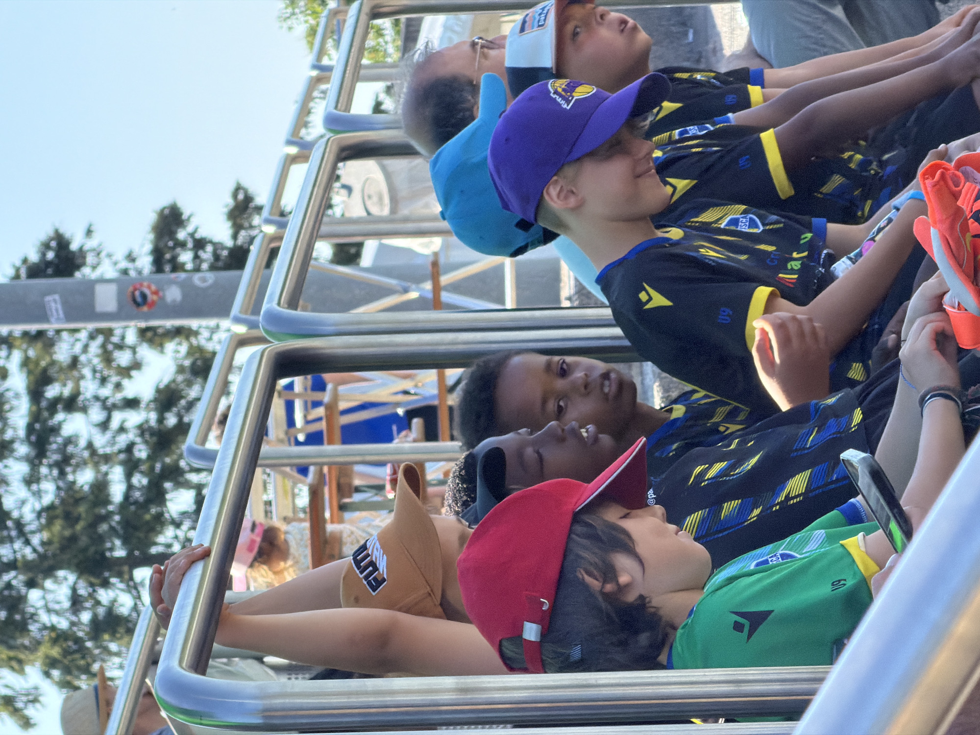 A group of children and adults are riding on an open-top tour bus in Fentange, Luxembourg. The bus is equipped with a chrome safety railing system. The riders are gazing upwards, presumably at a point of interest. The children are wearing colorful baseball caps and soccer jerseys, suggesting a team outing. The backdrop includes trees and a street sign.