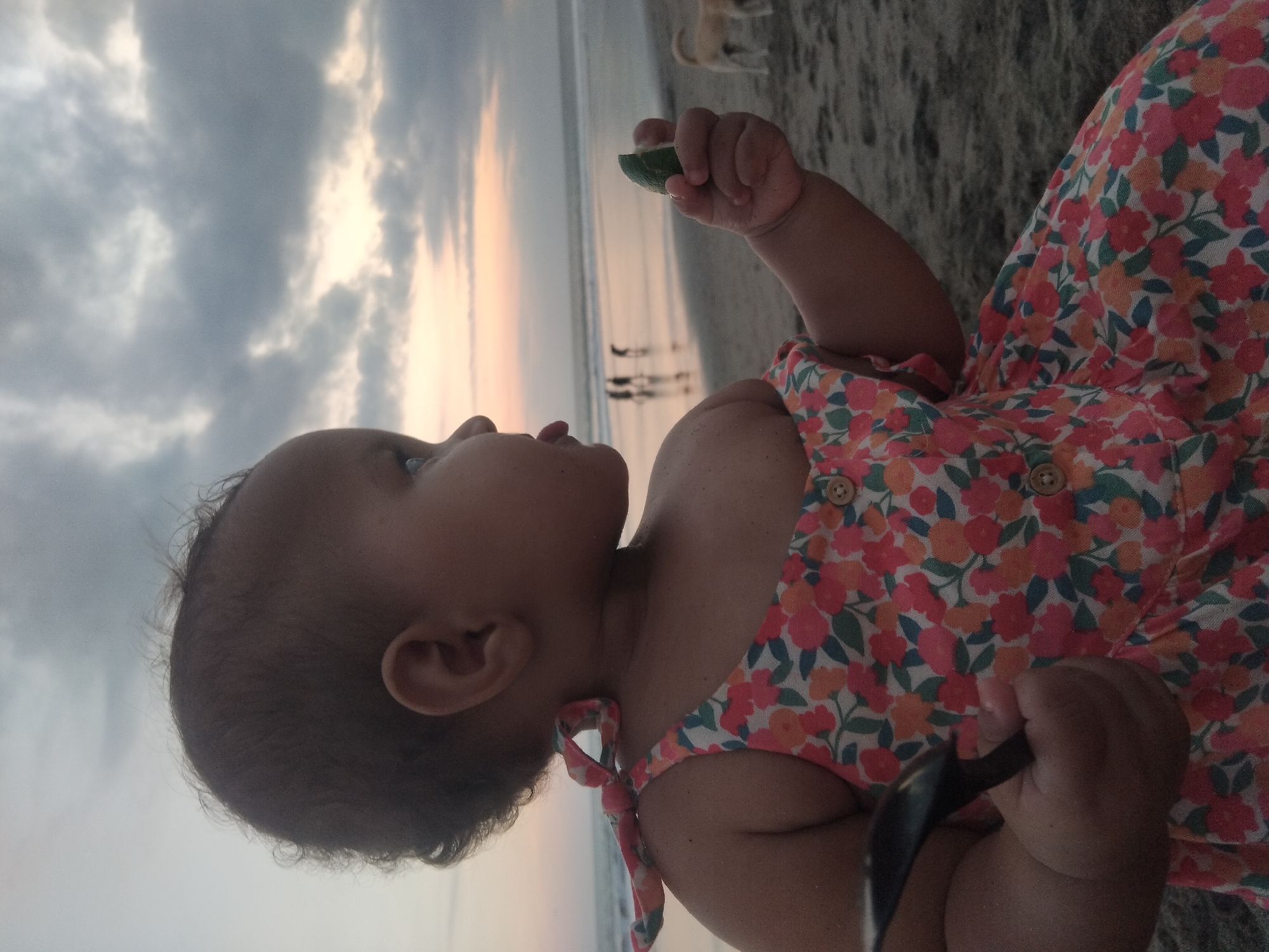 The image captures a close-up profile of a baby or toddler on a beach during either sunrise or sunset. The child, positioned on the right side of the frame, is looking upwards and towards the horizon on the left. The baby has dark hair and is wearing a vibrant floral dress featuring patterns of orange, pink, and green flowers with small brown buttons and a tie on the shoulder strap.
In the baby's right hand, a small, green wedge of fruit, possibly a lime, is held. The left hand clutches a dark, spoon-like object or toy. The baby's expression appears observant and curious as they gaze at the sky.
The background shows a vast expanse of sandy beach meeting the ocean. The sky displays a soft color gradient, transitioning from warm orange and pink tones near the horizon line to lighter gray and white clouds higher up. The lighting suggests the golden hour. In the distance, several blurry human figures are visible walking near the water's edge, along with a light-colored dog on the sand. The ocean reflects the sky's colors softly. There is no visible text in the image.