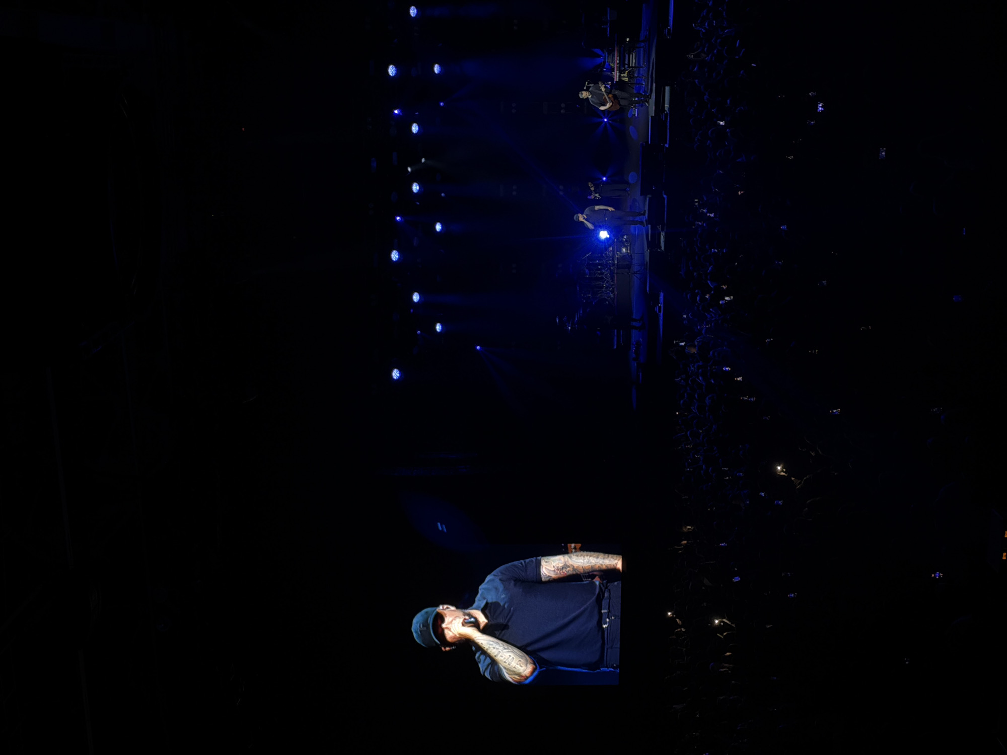 The image depicts a concert scene, possibly in Krasnoyarsk, Russia, given the user's request. A rapper with tattoos and a cap is performing on stage, holding a microphone. The stage is illuminated with blue spotlights. In the background, other musicians are present on a higher platform. A large crowd is visible on the right side, with many people holding up their phones, presumably to record the performance. The overall environment is dark, with the stage and performers as the primary sources of light.