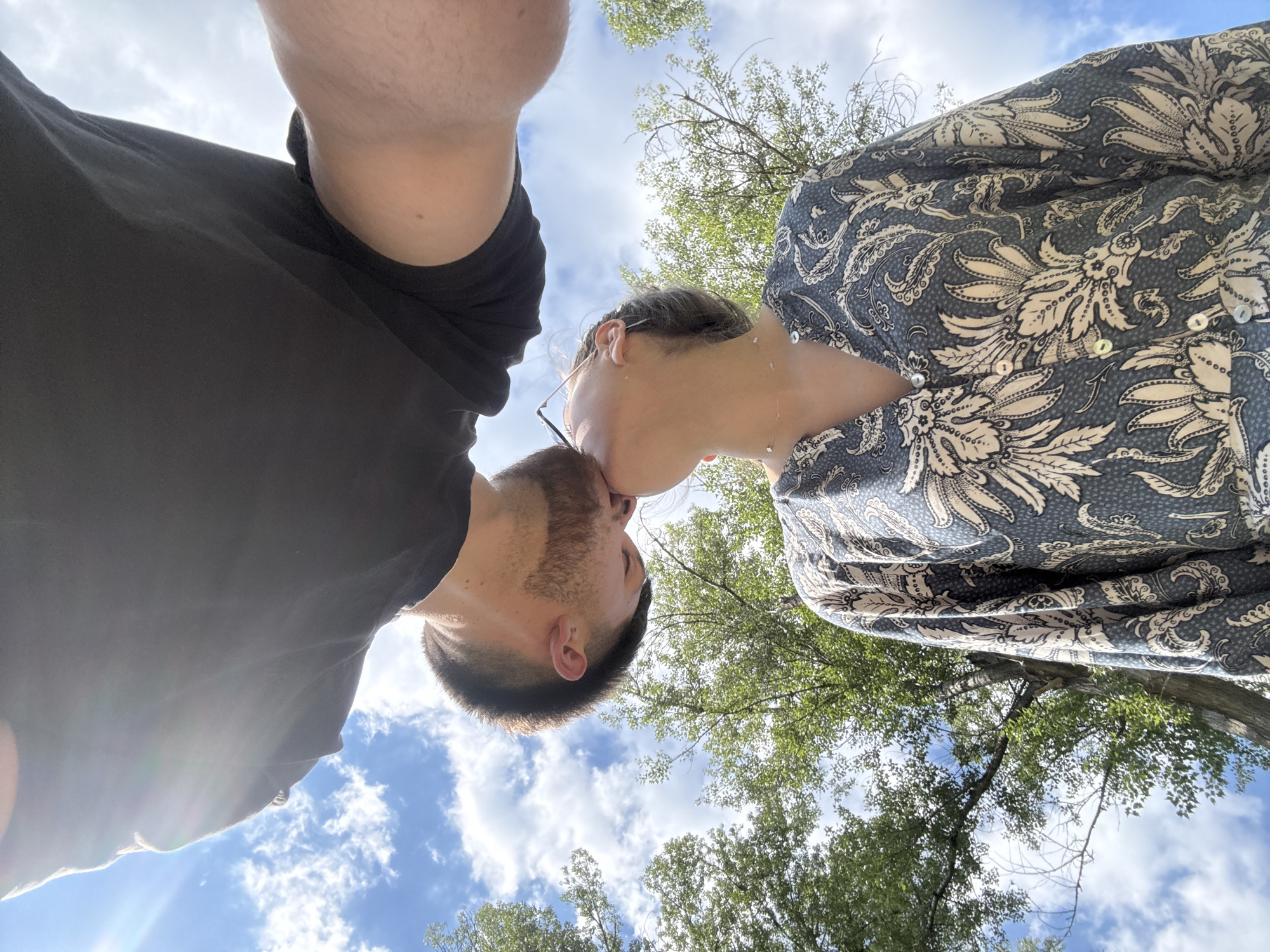 A low-angle shot captures two people kissing, with the sky and trees in the background. The person on the left has short, dark hair and a beard, wearing a dark t-shirt. The person on the right wears a patterned blouse and glasses. They are both facing upwards. The background shows a blue sky with white clouds and green treetops. There is no information to verify the location in the city of Blagoevgrad, Bulgaria.