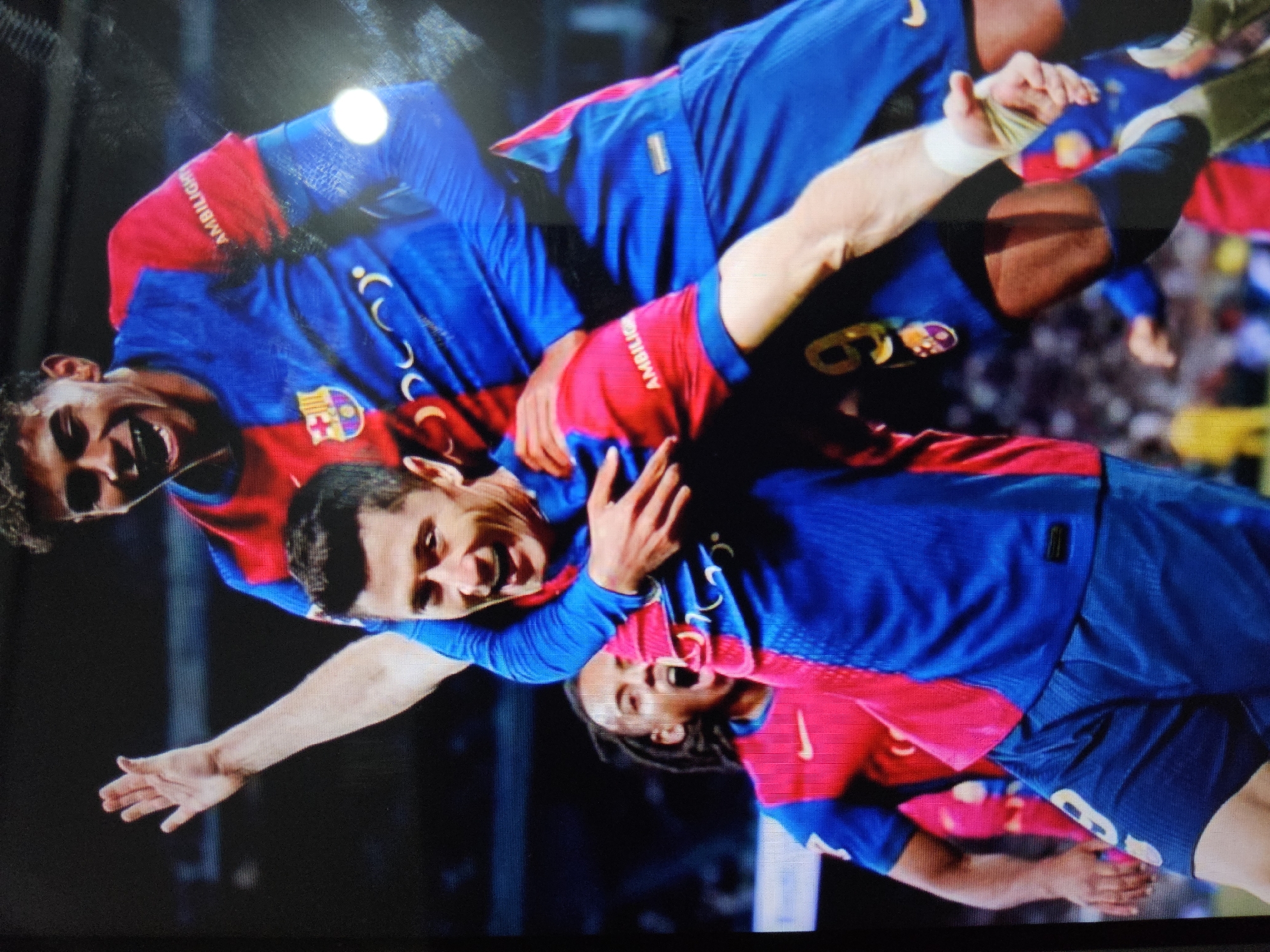 The image captures a moment of celebration between two male soccer players on a field. The player in the foreground, wearing a blue and red jersey and blue shorts, is being carried by another player. He has dark hair and is smiling broadly, his arms wrapped around the shoulders of the player carrying him. The player carrying him, also in a blue and red jersey, is looking upwards with a wide grin and has dark, curly hair. Both players' jerseys feature a crest on the left chest and a sponsor logo on the sleeve. In the background, other players in similar attire are visible, some with their mouths open in cheers. The environment appears to be a stadium with a blurred crowd in the distance. The image is a close-up shot, focusing on the players' joyous interaction. The location is not specified within the image itself, and therefore cannot be determined to be Payagaboh, Indonesia.