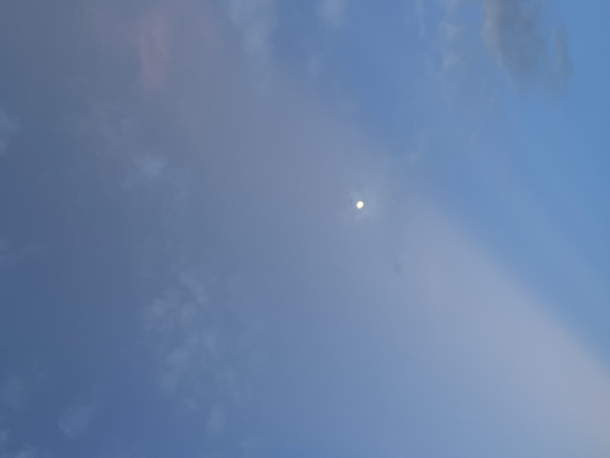 This media file is a photograph of the sky above Chiang Mai, Thailand. The sky is predominantly blue, with scattered clouds of varying opacity and texture. A bright, small object, likely the moon or a very bright star, is visible in the upper-middle portion of the frame, surrounded by a slight halo, suggesting it might be daytime or twilight. There is also a diffuse, vertical band of lighter, hazy light running from the lower right to the upper middle of the image, which could be caused by atmospheric conditions or light scattering. The overall impression is of a calm and possibly serene atmosphere, typical of a clear or partly cloudy sky. The lighting suggests it could be either late afternoon or early morning. No people, objects, text, or specific location cues beyond the general setting are visible. The image captures a natural scene without any discernible human activity or man-made structures.