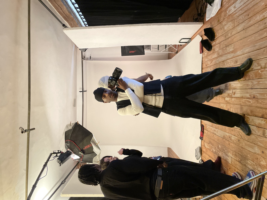 A man wearing a black beanie and a white and black sweater holds a camera in a studio setting. Behind him, another person with a white head covering appears to be observing. The studio features a white backdrop and a softbox light with a stand. In the foreground, two people dressed in black are partially visible. The floor is wooden, and there are various objects, including shoes and equipment, scattered around the edges of the scene.