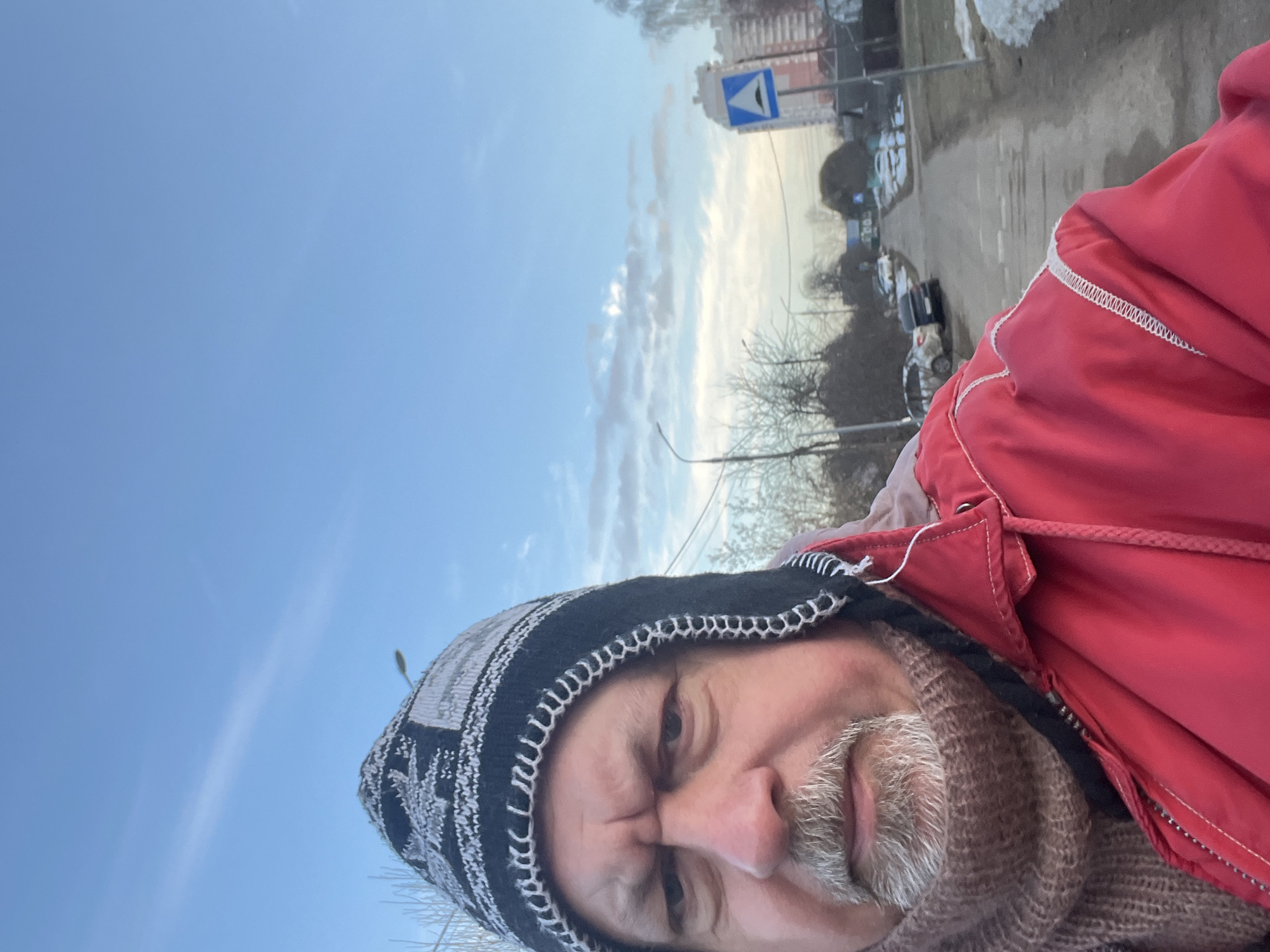 The image is a selfie-style photograph depicting a male individual outdoors. The man, seen from the chest up, has a grey goatee and mustache, and his brows are furrowed. He wears a black, grey, and white patterned knit beanie, a light brown textured knitted scarf, and a red zippered jacket with white decorative stitching on the shoulders.
The background shows a street environment under a partly cloudy blue sky. A narrow, unpaved or worn paved road extends into the distance, with bare deciduous trees and patches of melting snow visible along its sides. Several parked passenger vehicles, including a light-colored SUV and a dark sedan, are on the left side of the road. On the right, a blue triangular road sign with a white border and a black symbol indicating a speed bump is present. In the distance on the right, a multi-story light-colored residential apartment building is visible. This scene is located in the city of Podolsk, Russia.