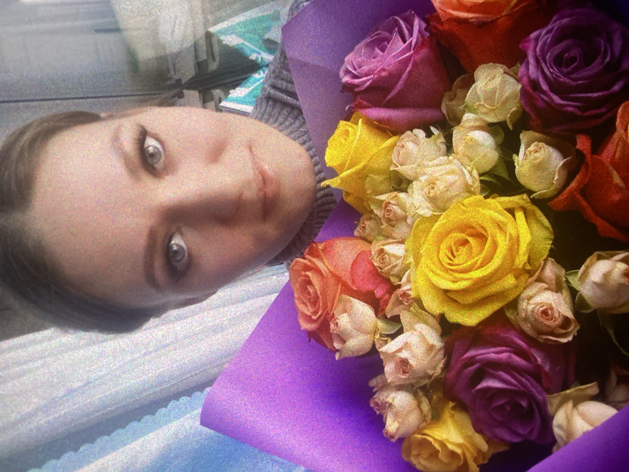 This rotated image (originally portrait) features a young woman holding a vibrant bouquet of roses. The woman's face is positioned on the upper left of the frame (when viewed upright), with her light eyes looking directly at the viewer and a gentle smile on her lips. She has light brown hair and is wearing a grey knitted sweater or top, visible on her left shoulder and arm.
The prominent object in the image is a large, colorful bouquet of roses, which occupies the right and lower-middle sections. The roses display a variety of hues, including deep purple, bright yellow, orange, and cream or light pink, with some smaller buds nestled among the larger blooms. The bouquet is wrapped in a distinct purple paper.
The background behind the woman and parts of the bouquet appears to be an indoor setting, with faint, blurry elements suggesting furniture or a room environment. There are light grey and bluish-white areas, possibly a wall or pleated blinds, creating a soft backdrop. The overall image has a noticeable grainy texture, characteristic of a high ISO photograph or a digital filter.
The activity depicted is likely a celebratory moment or a pose taken after receiving or arranging the flowers. The woman's expression suggests contentment or happiness. There is no visible text in the image.