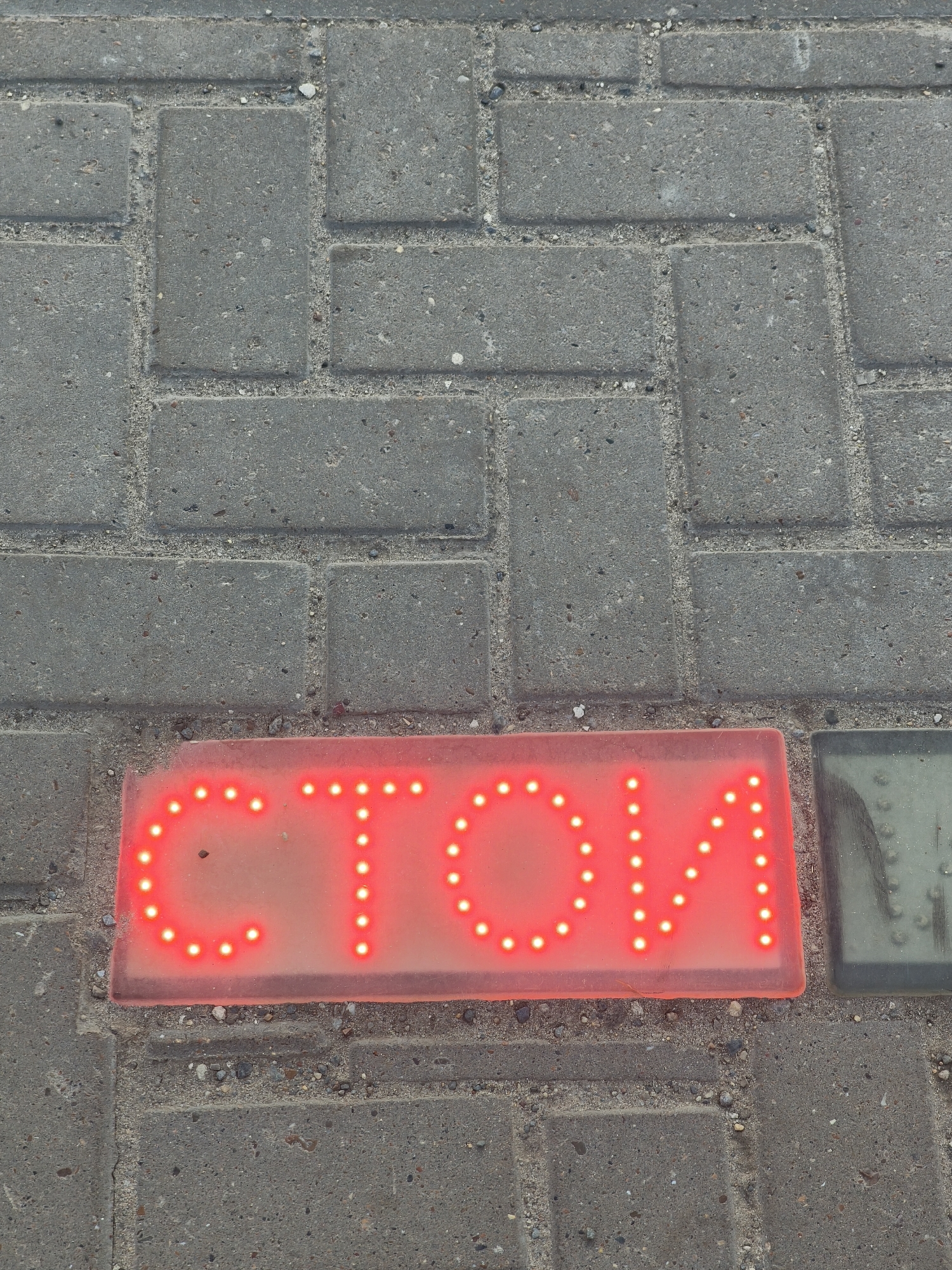 A red illuminated sign on a paved surface displays the word "СТОИМ" in Cyrillic letters. The sign is embedded in a sidewalk made of grey rectangular pavers. To the right of the red sign, a portion of a green square object is visible, also set into the sidewalk. The scene appears to be outdoors, likely in a public area. The word "СТОИМ" translates to "STOP" or "WE ARE STANDING" in English, suggesting a functional or informational purpose for the sign.