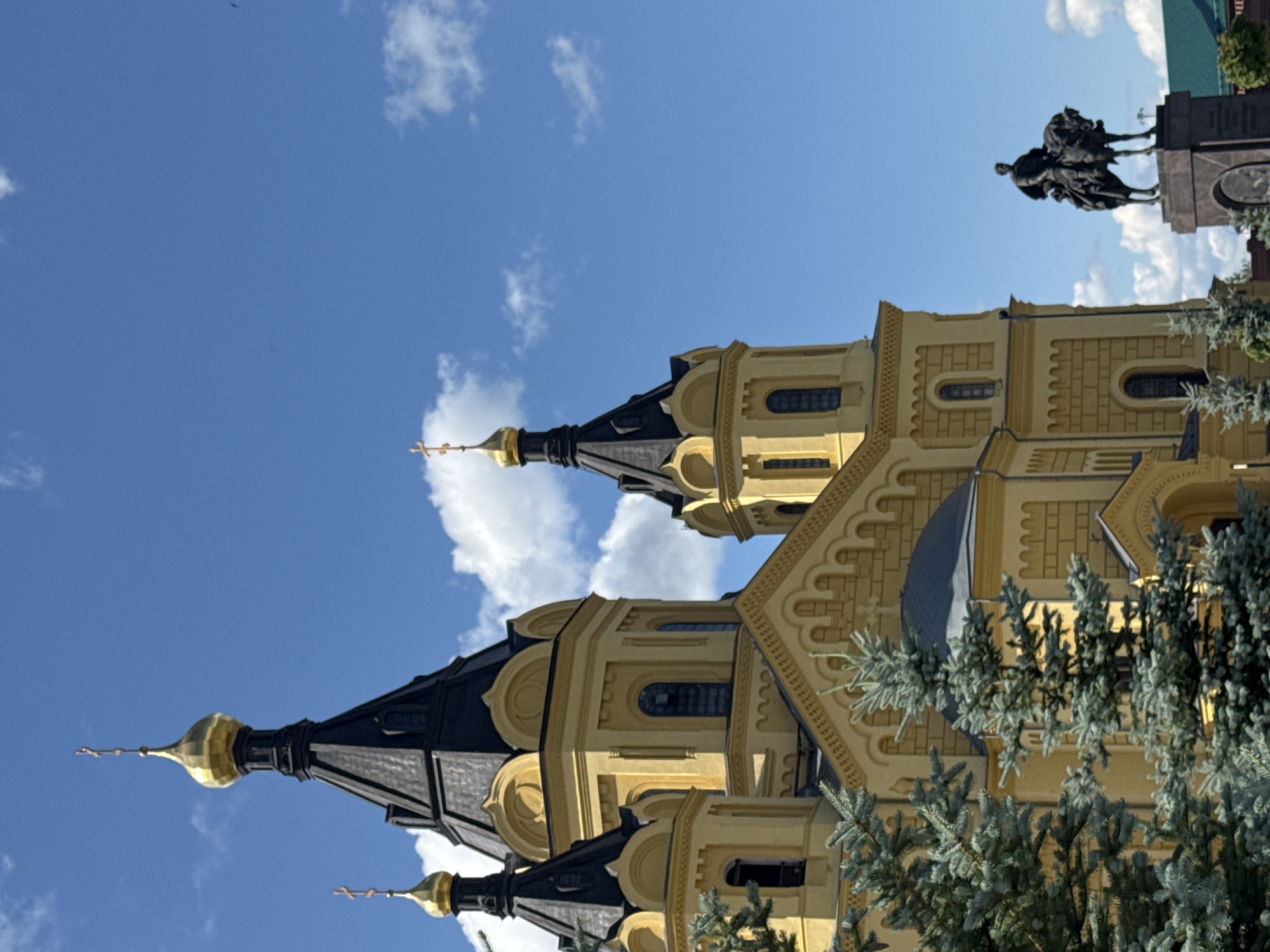 The image displays a portion of the Nizhny Novgorod Kremlin, a historic fortress overlooking the confluence of the Volga and Oka rivers in Nizhny Novgorod, Russia. The main subject is the Alexander Nevsky Cathedral, a prominent landmark within the Kremlin. The cathedral is characterized by its bright yellow facade, black tiled roofs, and golden onion domes topped with crosses. Several of its turrets and domes are visible against a bright blue sky dotted with white clouds. In the foreground, evergreen trees, likely spruce, partially obscure the lower part of the cathedral. To the right of the cathedral, a statue of a man on horseback is mounted on a pedestal. The statue is dark in color and appears to be made of bronze or similar material. The statue depicts a figure in historical attire, possibly a military leader or important historical figure. The overall scene suggests a sunny day with clear weather conditions, highlighting the architectural grandeur of the cathedral and the historical significance of the location.