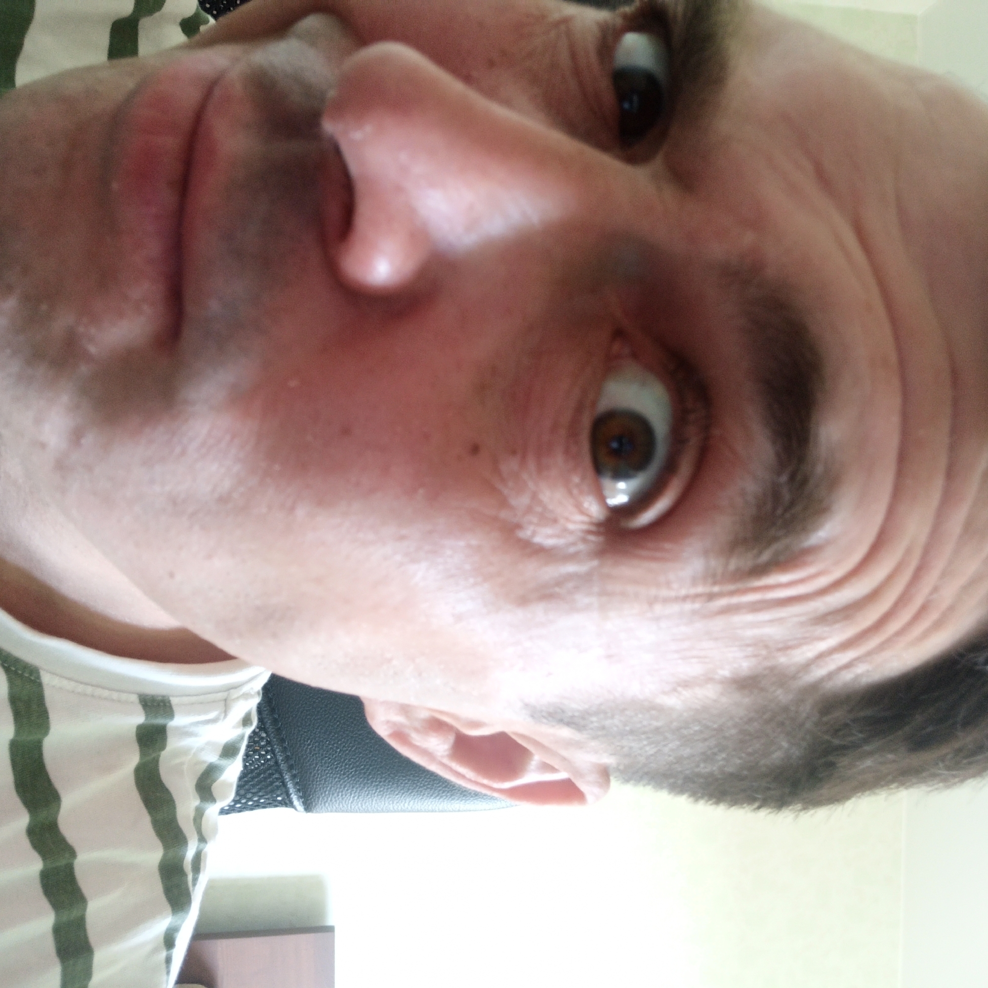 A close-up, Dutch angle, selfie of a man's face is presented. He has fair skin, dark hair, and brown eyes. His left eye is looking directly at the camera, while his right eye is slightly off to the side. His eyebrows are dark and arched. The man is wearing a striped t-shirt. The background is blurred, but appears to be an indoor setting with a light-colored wall and a dark chair. There is no indication of the specific location within Nizhny Novgorod, Russia.