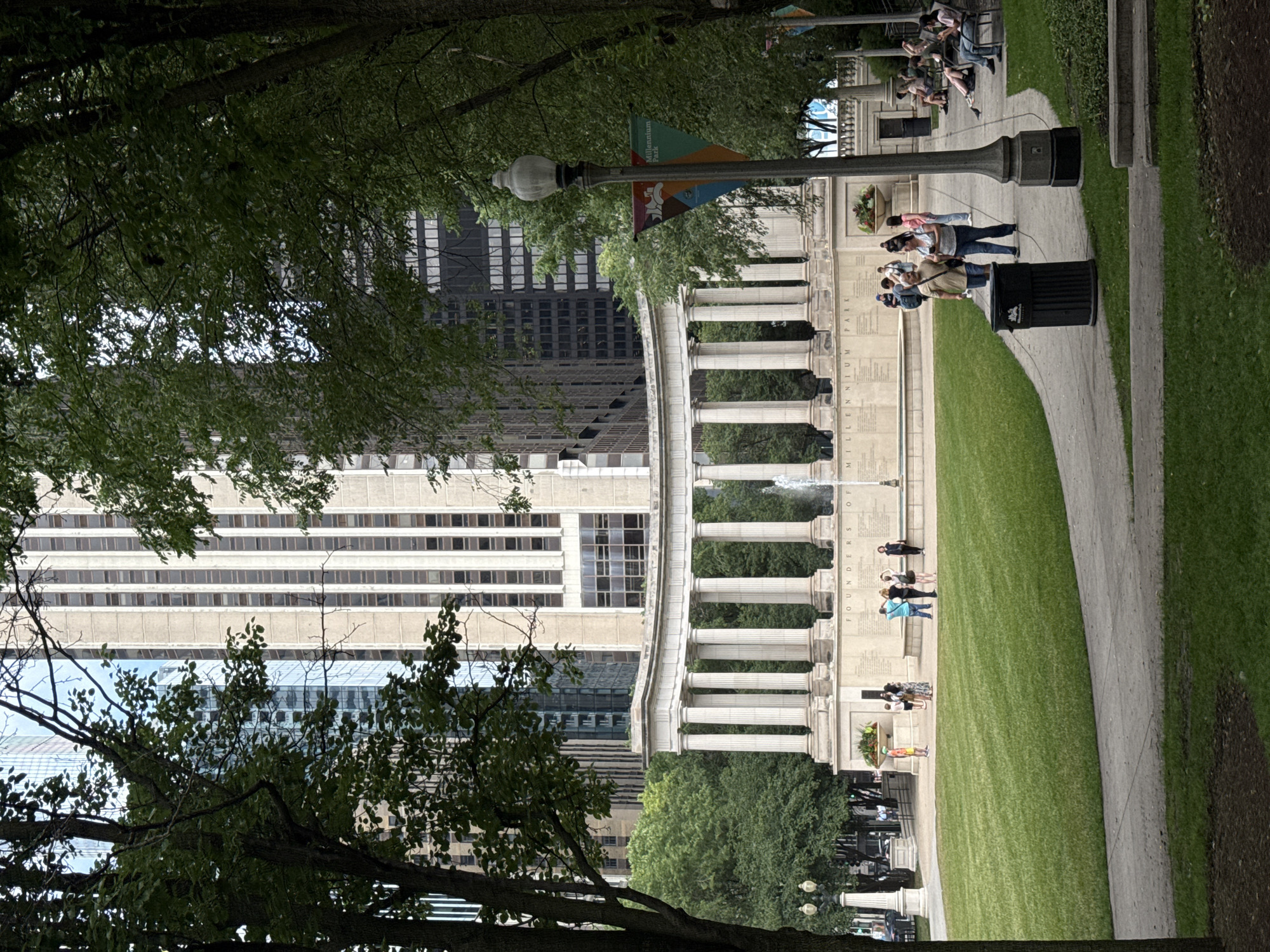 The image captures a scene in Millennium Park, Chicago, United States of America. In the background, a tall, modern skyscraper with a light-colored facade and numerous windows dominates the skyline. In front of the skyscraper, a semicircular colonnade with white, fluted columns stands prominently. Behind the colonnade, a fountain is visible, with water spraying upwards. The ground in front of the colonnade features text inscribed on a stone surface, partially obscured by a planter box.
In the mid-ground, a large, well-maintained green lawn extends towards the foreground. A paved pathway curves through the lawn, with a black trash receptacle located next to it. Several people are scattered across the park. On the lawn in front of the colonnade, a group of individuals are gathered, some appearing to be conversing or looking at the monument. Further back, near the pathway and on benches, more people are seated or walking. A lamppost with a decorative top stands to the right of the pathway, adorned with a banner that reads "Millennium Park." Trees with lush green foliage frame the scene on both the left and right sides, with branches and leaves obscuring parts of the background buildings and structures. The overall environment suggests a public park setting on a cloudy or overcast day.