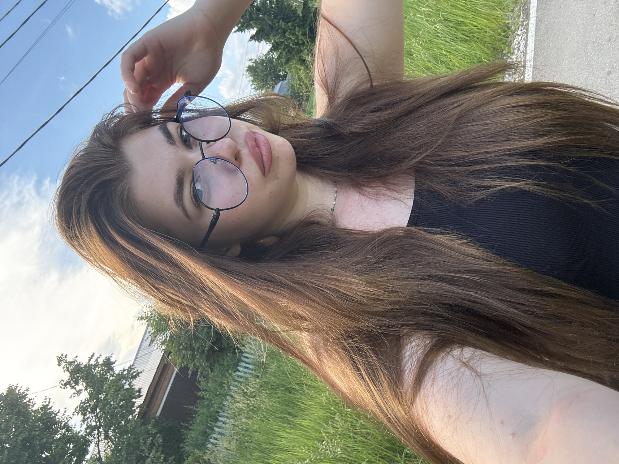 The image shows a young woman with long brown hair, wearing glasses and a black tank top, standing outdoors. She is looking at the camera and her right hand is raised to her head. The background consists of a rural setting with green grass, trees, a white fence, and a building. The sky is blue with some clouds. There are also power lines visible in the upper part of the image. The location is identified as Мещёры, Russia.