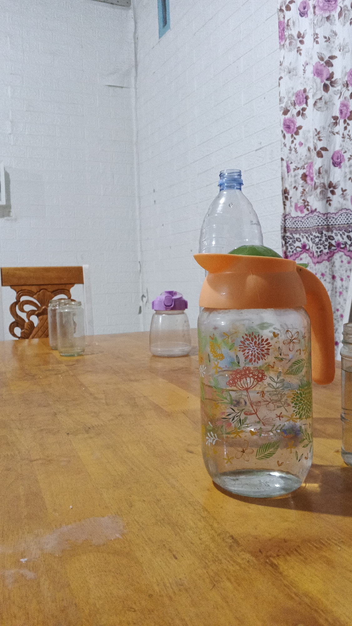 The image depicts a wooden table with several objects on it, including a glass pitcher decorated with floral patterns, a clear plastic bottle placed on top of the pitcher, a small glass jar with a purple lid, and two other clear glass jars. A wooden chair with intricate carvings is partially visible on the left side of the frame. In the background, a white brick wall and a curtain with a floral design are present. The scene appears to be indoors, possibly a home or a dining area, and is located in Kampung Rempak, Indonesia.