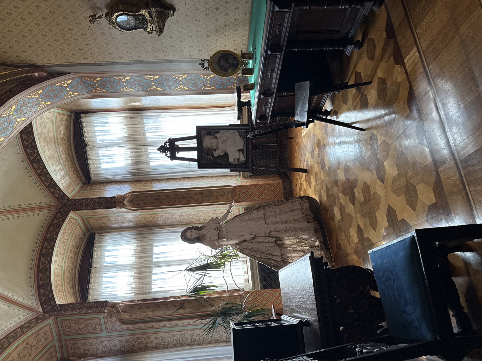 The image depicts an ornate room in Wernigerode Castle, Germany. The room features a parquet floor with a geometric pattern. In the center of the room stands a life-sized cardboard cutout of a woman in a voluminous 19th-century dress. To the left, a dark wooden desk with carved details is visible, accompanied by a dark wooden chair with a blue upholstered seat. To the right, a larger dark wooden desk with a green surface holds several framed photographs and a decorative clock. An easel displays a framed portrait of a baby. The walls are adorned with patterned wallpaper and decorative painted arches above tall windows covered by sheer white curtains. A large potted plant is situated near the cutout figure. The room is illuminated by natural light filtering through the windows.