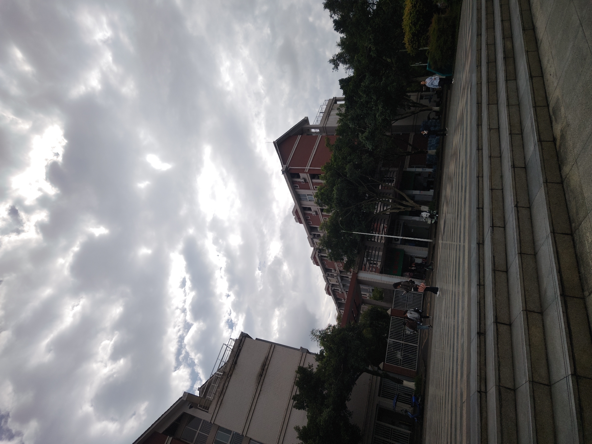 The image depicts an outdoor scene in Chongqing High-tech Zone, China. Two buildings, one brick and the other beige, are visible under a cloudy sky. Trees partially obscure the buildings. People are walking along a paved area with stairs.