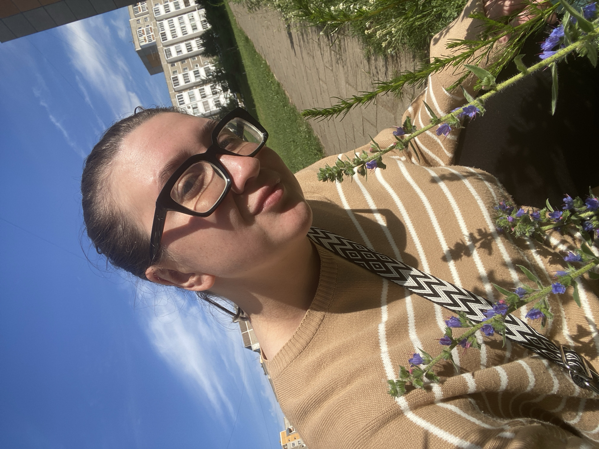 A medium shot selfie shows a young woman wearing glasses and a beige and white striped sweater, holding a small bouquet of purple wildflowers in front of her. She is outdoors, with a bright blue sky and wispy clouds above. Behind her, apartment buildings and green grass suggest an urban park or residential area. The woman's expression is a gentle smile. The focus is on her face and the flowers, with the background slightly blurred. The image appears to be taken on a sunny day.