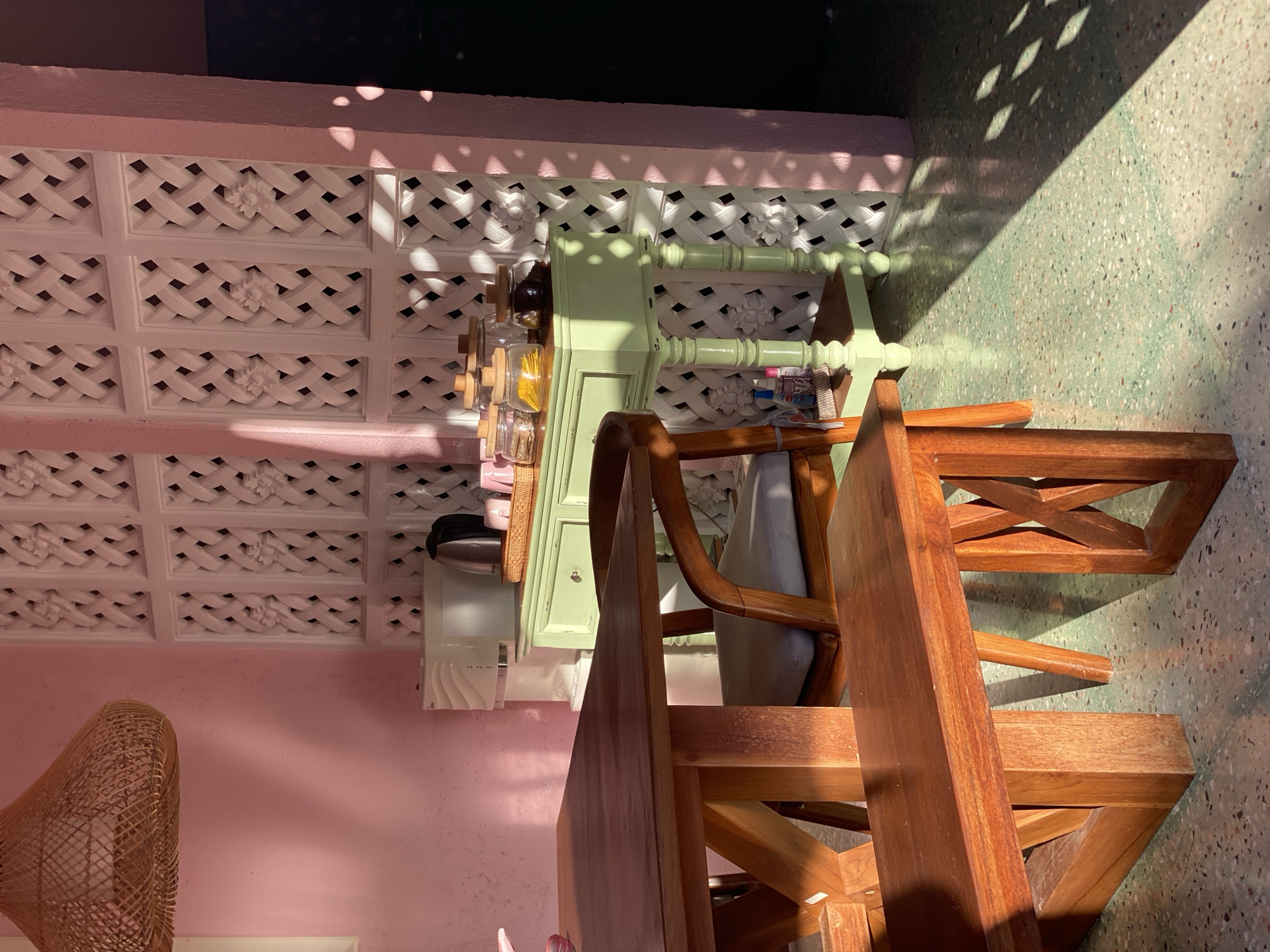 This image captures an indoor scene with a wooden dining table and bench in the foreground, bathed in sunlight. Behind the table, a light green console table holds several glass jars containing what appears to be dried goods or spices, and a wicker basket with a few pink items. A white appliance, possibly a water dispenser, is partially visible behind the console table. The wall behind the console table is decorated with white, lattice-like panels featuring floral motifs. The room is painted a soft pink. The floor is a speckled green terrazzo. Sunlight streams in from the left, casting distinct shadows from the lattice panels onto the wall and floor, creating a patterned effect. A woven lampshade hangs from the ceiling in the upper left corner. The scene is static and devoid of any people. The overall ambiance is one of bright, warm, and possibly domestic tranquility.