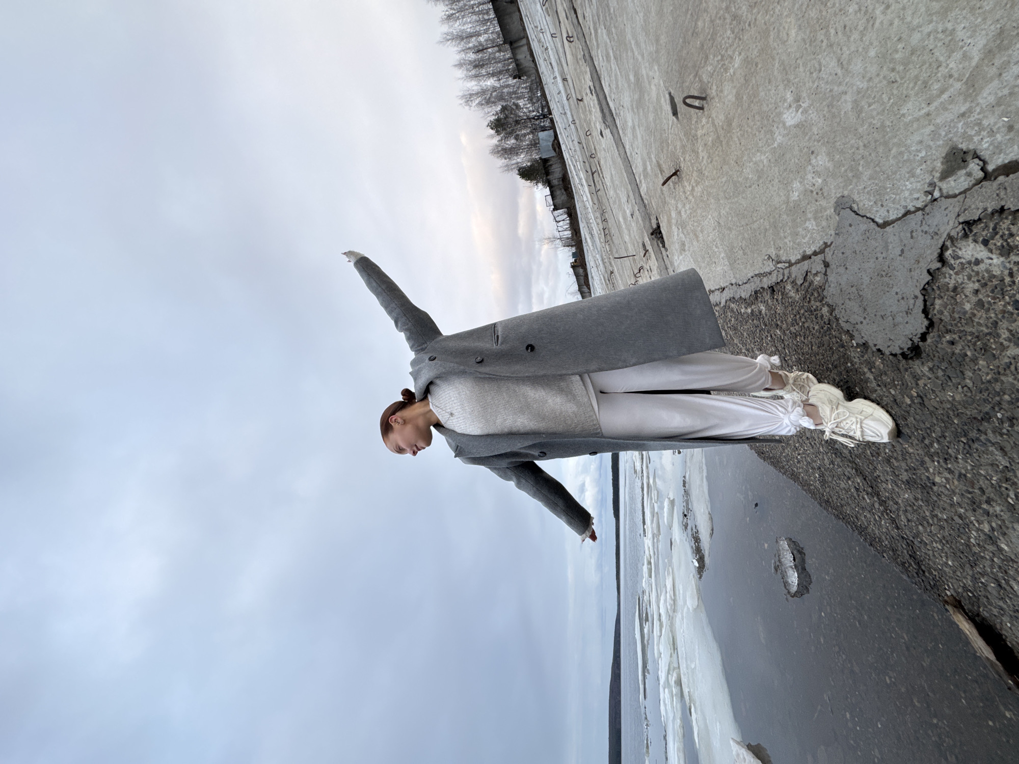 A full-body, horizontally oriented shot depicts a young woman standing on a weathered concrete embankment next to a body of water. The woman, shown in profile, is positioned on the right side of the frame, facing left. She wears a long, light grey overcoat, a white sweater, white pants, and white chunky athletic shoes. Her brown hair is pulled back, and her arms are outstretched, one pointing upwards and slightly backward, the other downwards and slightly forward, as if balancing or mimicking flight. She is looking towards the horizon.
The setting appears to be an industrial or natural waterside location, possibly in Chaikovsky, Russia, given the context. The concrete embankment is cracked and shows signs of wear, with rebar visible in places. To the woman's left, the concrete surface continues into the background, where bare trees and some distant structures can be seen under an overcast sky. To her right, the body of water is partially frozen, with large pieces of ice floating or accumulated near the shore. The sky is a uniform, light grey, indicating an overcast day. The overall impression is one of cold weather, likely winter or early spring, and a somewhat desolate, open landscape. There is no visible text.