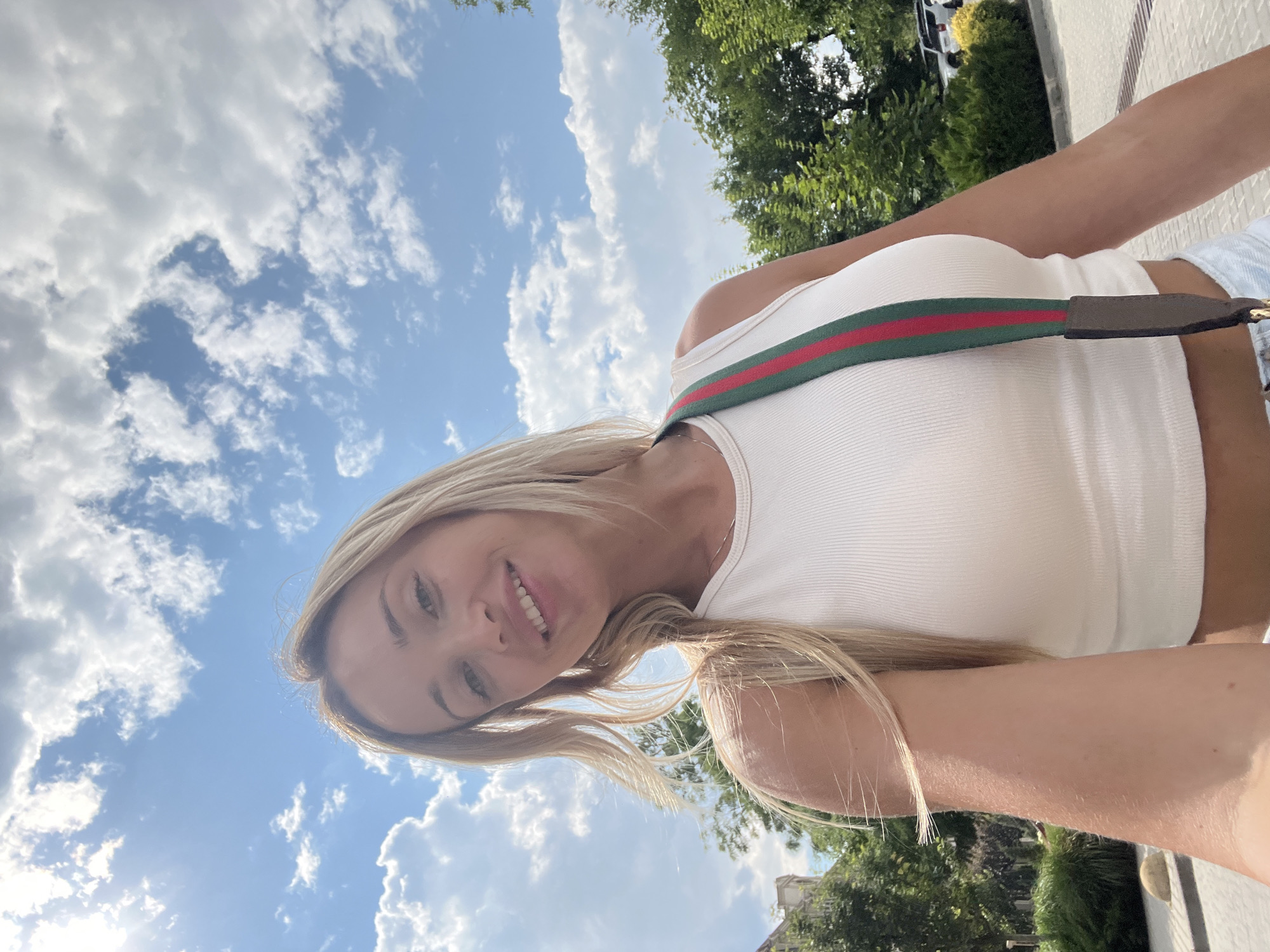 A young woman with blonde hair smiles at the camera as she stands outdoors. She wears a white ribbed tank top and has a strap with red and green stripes draped over her shoulder. The sky is blue with scattered white clouds. Trees and some buildings are visible in the background, suggesting an urban park or street setting. The image appears to be a selfie taken on a sunny day.