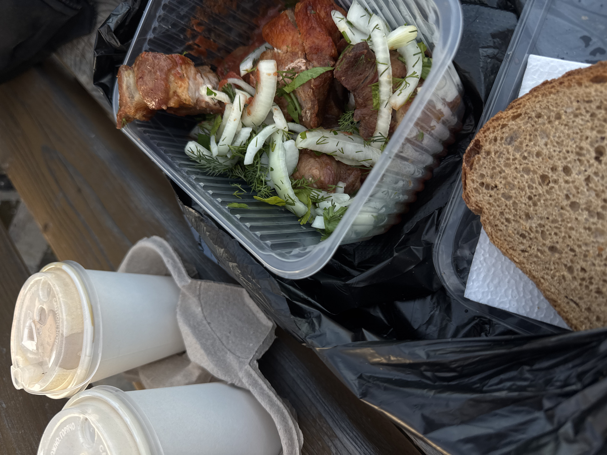 The image depicts a take-out meal arranged on a wooden surface, likely a bench or table, in the city of Chaikovsky, Russia. On the upper left, two white disposable beverage cups with clear plastic lids are secured in a grey cardboard carrier; the rightmost cup contains a light brown liquid, possibly a coffee-based drink. A prominent black plastic bag covers the lower left and extends partially to the right. In the center-right, a clear rectangular plastic container holds multiple pieces of cooked meat, possibly shashlik, which are reddish-brown and appear grilled or roasted. The meat is topped with generous slices of white onion and fresh green herbs, including dill and possibly parsley. A reddish liquid or sauce is visible at the bottom of the container. In the lower right, a single slice of dark, porous bread, likely rye bread, rests on a small white textured napkin, positioned near another part of the black plastic bag. No individuals are visible in the scene.