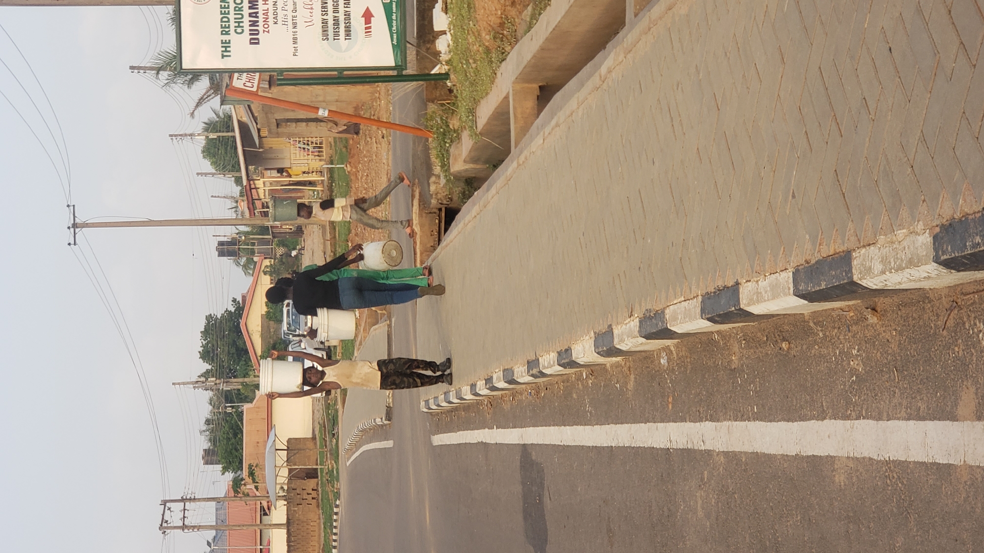 The image shows a road scene, likely in Narayi, Nigeria, based on the sign for "The Redeemed Church of God Dunamis Zonal Headquarters Kaduna," and the general environment. The image is rotated. Three individuals are visible on the right side of the road, close to a retaining wall. The first man on the left of the frame is carrying a white bucket on his head, his arms raised. The second person wears a black top and is carrying a white bucket on their head. The third person appears to be carrying a white bucket on their head as well. A fourth man further down the road is also carrying a white bucket. The road itself has a white line marking. There are power lines and buildings in the background.