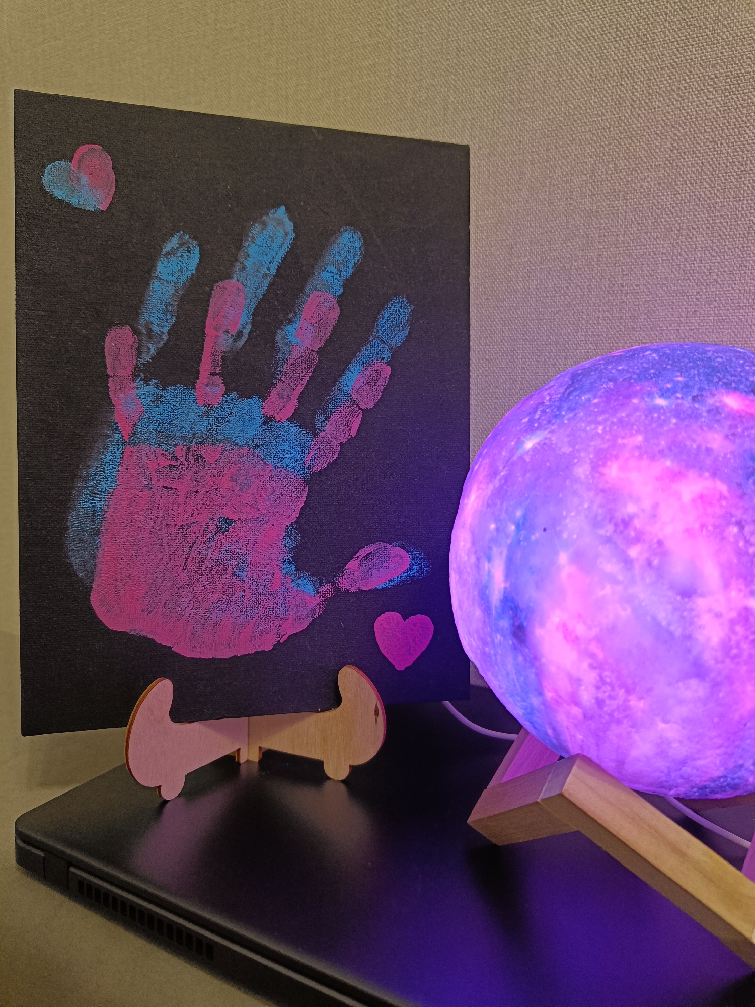 A close-up, medium shot captures a piece of art and a decorative lamp. The artwork, a black canvas adorned with pink and blue handprints, is propped up by a light-colored wooden easel. Two small heart shapes, one pink and one blue, are also visible on the canvas. To the right of the artwork is a spherical lamp, emitting a soft purple and pink glow, supported by a wooden stand. The lamp rests on a dark surface, possibly a laptop. The background consists of a textured, light-colored wall. The scene appears to be an indoor setting, possibly a home or office. The image does not contain any information that would allow for its location to be identified in Krasnodar, Russia.