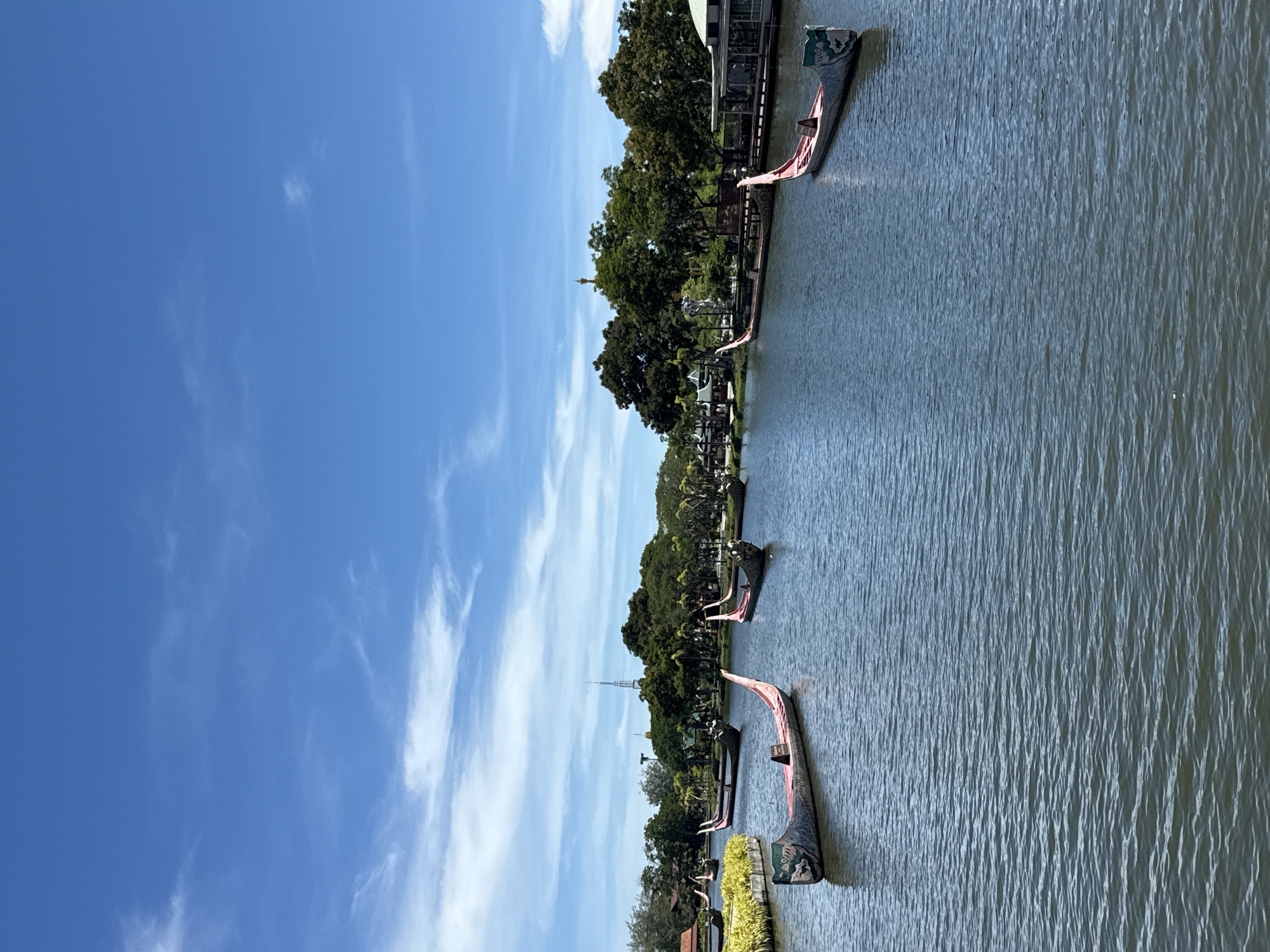 The image depicts a waterway in Bang Pu Subdistrict Municipality, Thailand, with several longboats docked along the bank. The sky is a bright blue with wispy clouds. The water has a rippled surface. Trees line the bank, and a structure is visible behind them. A tall spire-like structure can be seen in the distance behind the trees.