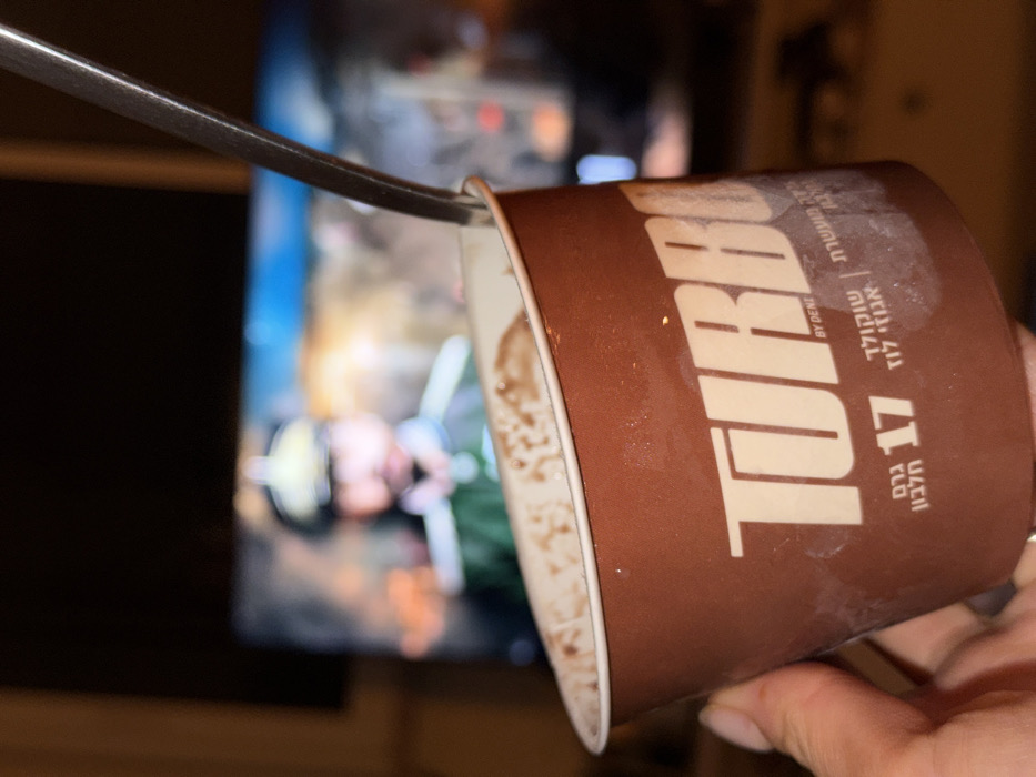 The image displays a close-up, angled view of a person's left hand holding a brown plastic cup, against a blurred background of a television screen in a dimly lit room.
**Objects and Subjects:**
The central object is a cylindrical brown plastic cup, branded with "TURBO" in large white capital letters and "BY DENE" below it. Hebrew text on the cup identifies the contents as "שוקולד אגוז לוז" (Chocolate Hazelnut) and specifies "17 גרם חלבון" (17 grams protein). The cup contains a partially consumed dark brown, viscous substance, with residue visible along the inner rim. A silver metallic spoon is inserted into the substance, angled upwards. The cup is held by a light-skinned hand, with a metallic ring visible on the ring finger.
In the blurred background, a television screen is visible, displaying a scene that features a blurred male figure. This figure has a mustache and is wearing a green military-style uniform with a high collar and a distinctive, possibly pointed, helmet. The background on the screen appears to be an outdoor setting with blurred elements of blue, orange, and brown.
**Actions and Environment:**
The scene captures the action of an individual consuming a protein dessert while watching television. The setting is an indoor, likely residential, environment characterized by low ambient lighting, with the television screen serving as the primary light source in the background.
**Location:**
Based on the visible Hebrew text and the brand "TURBO BY DENE," which is an Israeli food product (produced by Tnuva), the scene is definitively located within Israel. While the specific city of Tel-Aviv is not visually explicit, the presence of a common Israeli consumer item in a residential context is consistent with such a location within a major Israeli city.