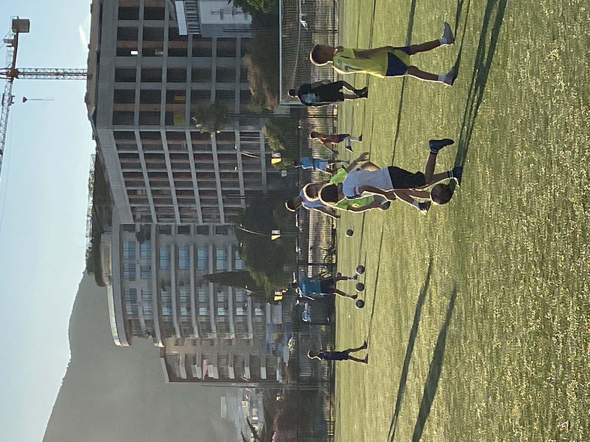 This image depicts a group of young boys practicing soccer on an artificial turf field in Budva, Montenegro. In the background, modern apartment buildings and a construction crane are visible, suggesting an urban or semi-urban setting. The sun is setting, casting long shadows across the field and illuminating the scene with a warm, golden light. Several boys are actively engaged in training drills, with some dribbling soccer balls and others standing around. One adult male, possibly a coach, is also present on the field. The overall atmosphere is one of athletic activity and recreation, set against the backdrop of a developing coastal city.