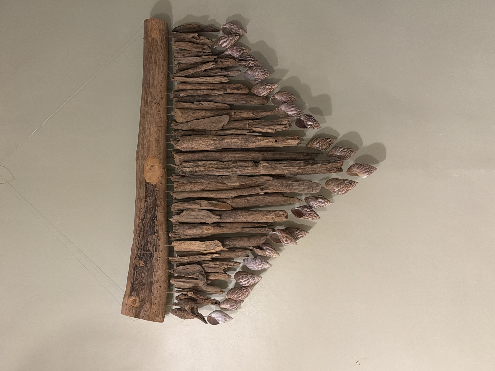 The image shows an art piece, possibly a wall hanging, composed of driftwood and seashells arranged in a cascading pattern. The driftwood pieces are horizontally aligned, decreasing in length from top to bottom, and are held together by what appears to be thin wire or string. Each row of driftwood is paired with a seashell.
The background is a flat, light green wall.
Although the prompt mentions North Kuta, Indonesia, there is no visual information within the image that confirms this location.