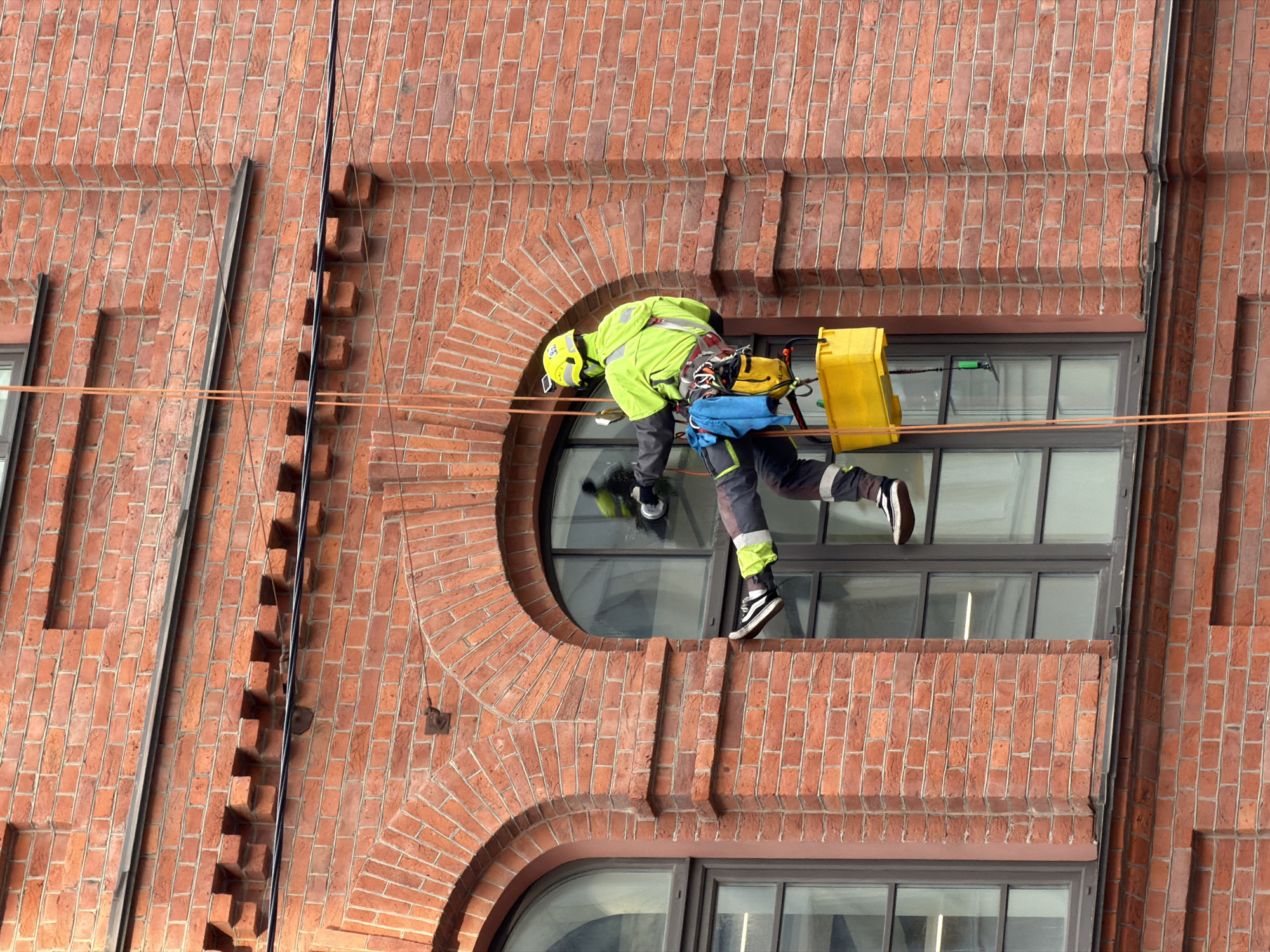A worker performs maintenance or window cleaning on the exterior of a historic red brick building in Saint Petersburg, Russia. The individual is suspended by ropes, utilizing rope access techniques, and is seen leaning into a large arched window opening. They wear a bright, high-visibility lime green jacket and helmet, dark pants with reflective stripes, and black and white sneakers. A safety harness and various ropes secure the worker, and a yellow bucket or container is attached to their equipment, likely holding cleaning supplies. A cleaning tool with a green handle is visible, reflected in the windowpane.
The building's facade is constructed from distinctive red brick, featuring intricate architectural details such as decorative arches, cornices, and recessed brickwork patterns. The scene is brightly lit, suggesting clear daytime weather conditions. No visible text is present in the image.
