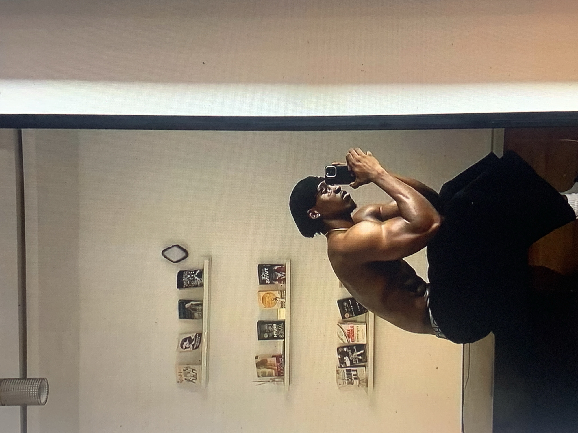A shirtless male individual, appearing to be of Black ethnicity with a muscular physique, is captured in a mirror selfie within an indoor setting in the city of Unknown, Unknown. The individual is wearing a black durag, eyeglasses, and black shorts, revealing a visible light-colored waistband. He is seated on a dark, low-profile surface. He holds a black smartphone with both hands, angling it towards the mirror to capture his reflection, with his gaze directed towards the phone screen.
The background consists of a plain white wall adorned with three white floating shelves. From top to bottom, the shelves display various small rectangular objects, primarily books, with some text appearing reversed due to the mirror reflection.
- The uppermost shelf contains four books and a small, dark, irregularly shaped decorative mirror to its right.
- The middle shelf holds four books, including one with partially visible inverted text "TRUTH" and another with inverted text "ICE TWINS." A white reed diffuser is positioned to the left of this shelf.
- The lowest shelf displays four books.
A cylindrical silver light fixture with a mesh texture hangs from the ceiling in the upper left corner of the scene. The reflection on the right side of the image is bounded by a black mirror frame, beyond which a bright vertical strip is visible. The floor reflected beneath the seated individual appears to be a light-colored wood or tile. The lighting is bright, illuminating the scene and highlighting the individual's skin.