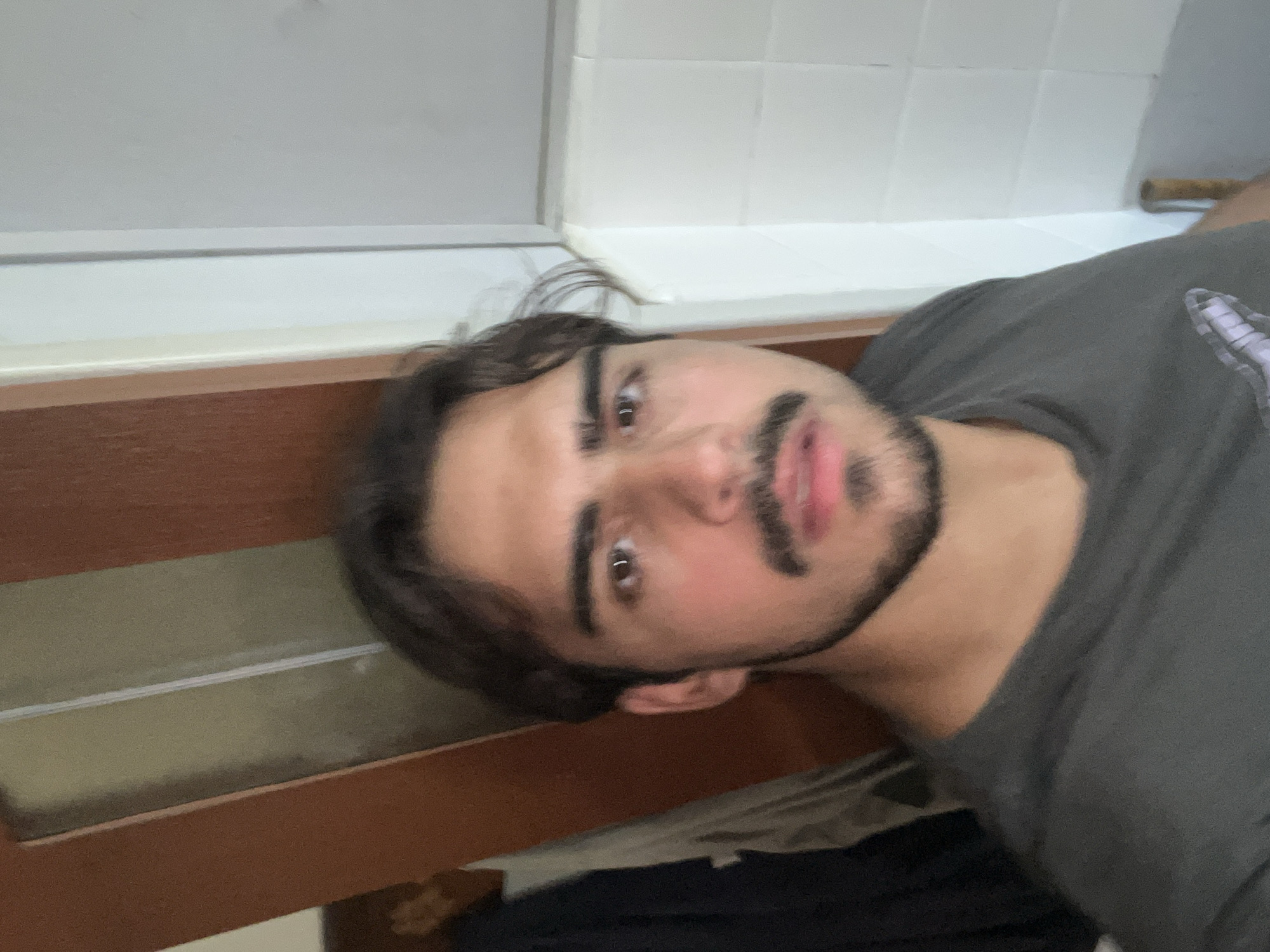 A close-up, angled selfie shows a person with brown hair, brown eyes, and a mustache. The person is wearing a grey t-shirt. The background features a tiled wall and a wooden frame structure, possibly a window or a counter. There are no details in the image to indicate the specific location in Buenos Aires, Argentina.
