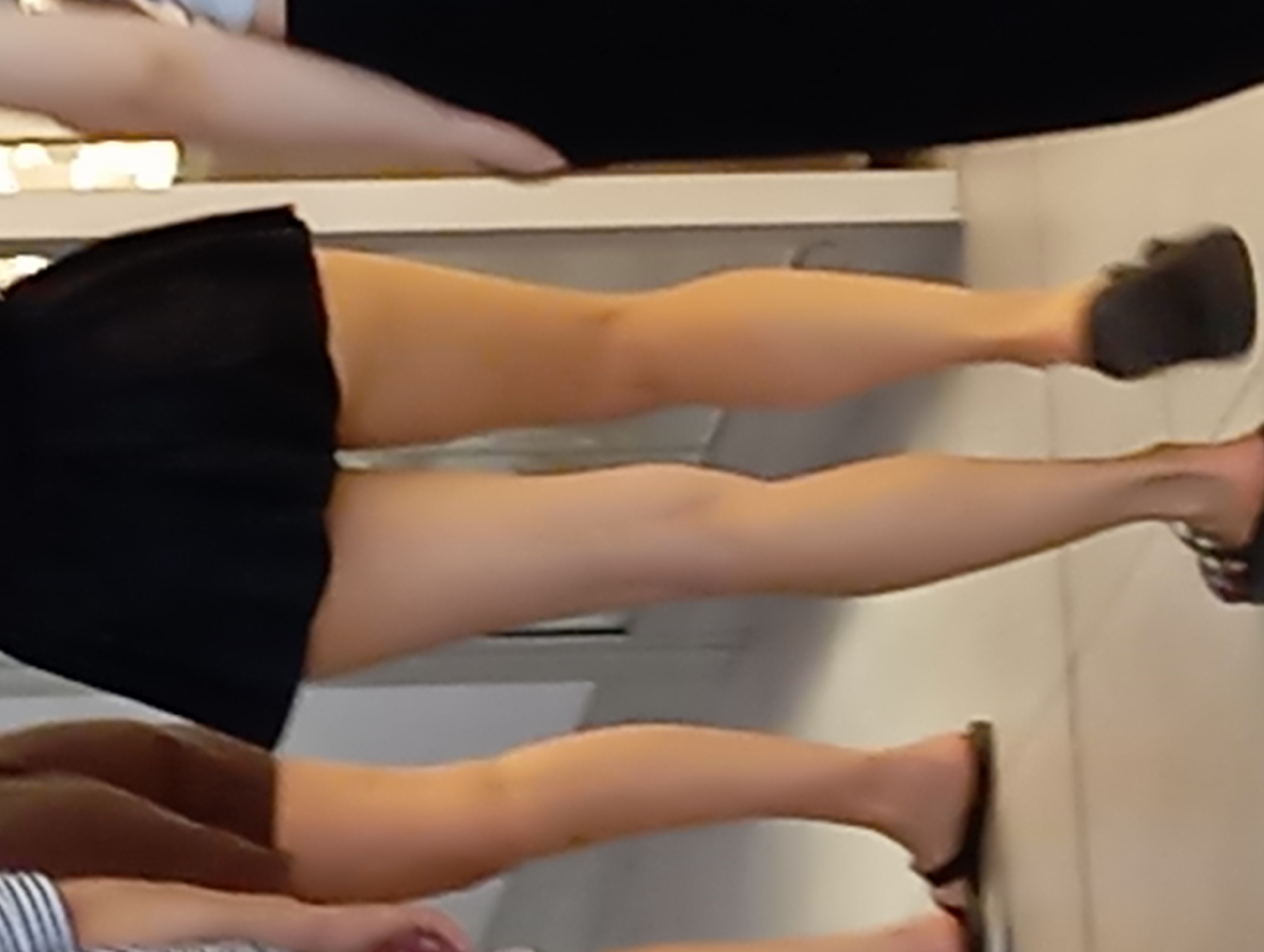 A rear-view image shows the legs of three individuals walking. The person in the center is wearing a short black skirt and dark sandals. To their left, another person wears brown shorts and dark sandals. To the right, a third person wears a black garment and dark sandals. They are walking on a light-colored tiled floor indoors, possibly in a commercial establishment in Nitra, Slovakia. The background is blurred and appears to be a wall with some lighting fixtures.