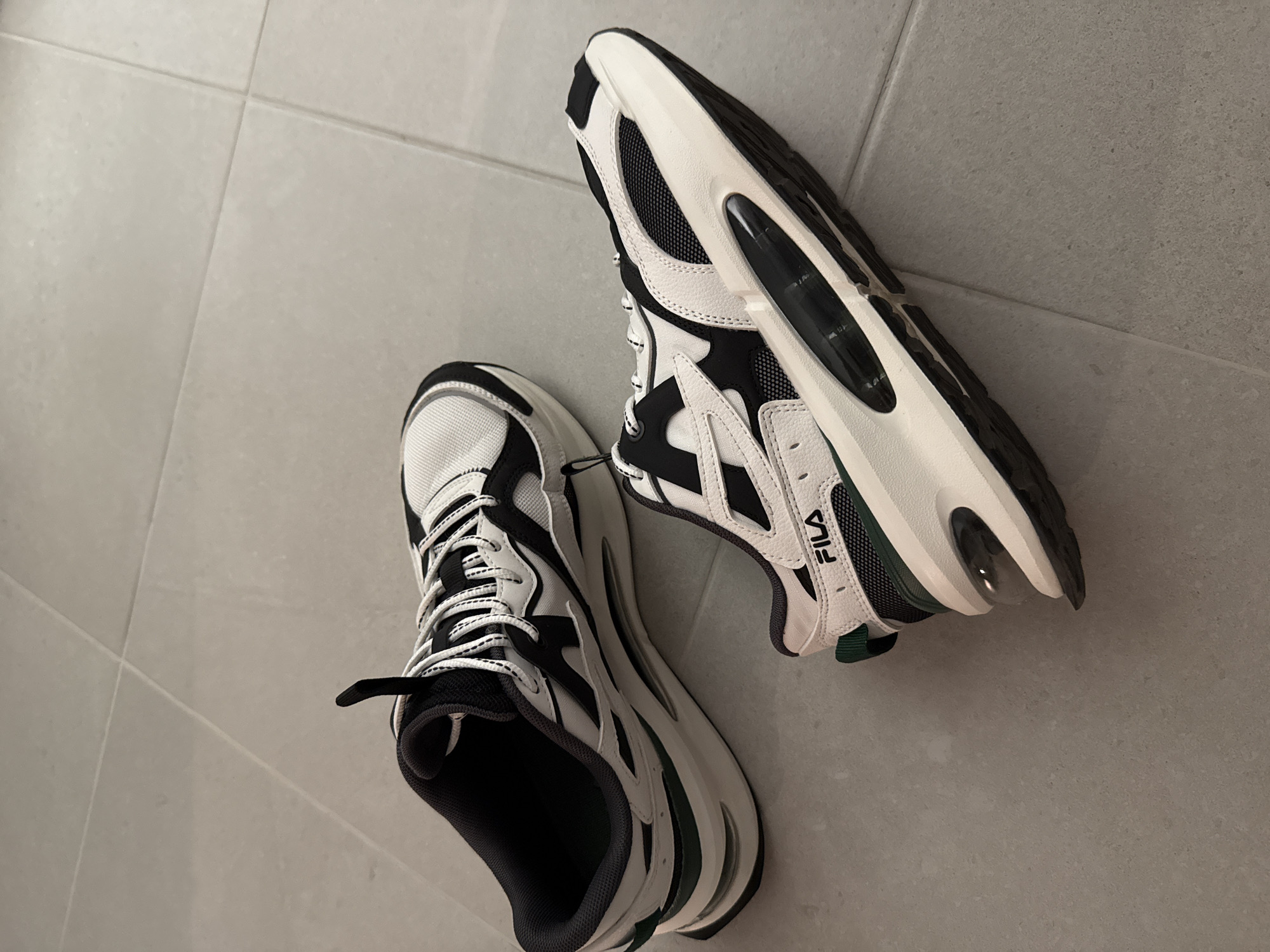 Two black and white Fila athletic shoes are displayed on a light gray tiled floor. One shoe is positioned on its side, revealing the interior lining, while the other shoe is laid flat, showcasing its sole and the "FILA" logo on its side. The shoes appear to be new or in excellent condition, with no visible signs of wear or damage. The lighting is bright and even, suggesting an indoor setting, possibly a retail store or a home. There are no people or any other objects visible in the scene. The location is unknown and not specified.
