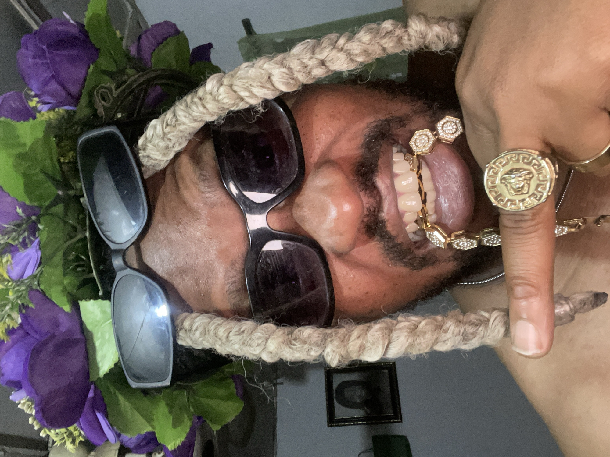 The image shows a person's face close-up, oriented upside down in the frame. The person is wearing sunglasses and has a rope-like headpiece adorned with purple flowers. The person's tongue is protruding, displaying a gold and jeweled grill. A large gold ring with a Medusa-like design is visible on their finger. The person is in an indoor environment. The location is not evident from the image.