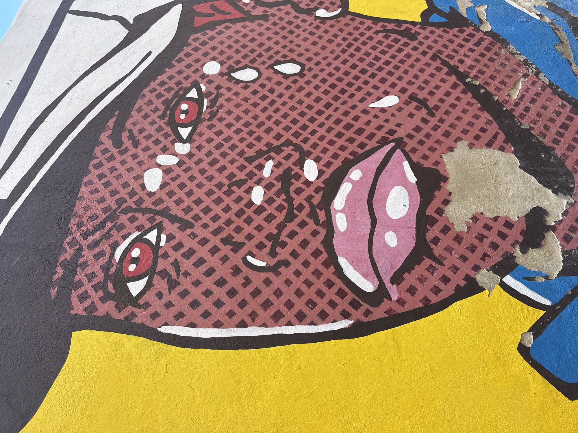 The image is a close-up section of a Pop Art-style mural, possibly depicting a female face in distress, given the eyes with red pupils and white highlights suggestive of tears. The face is rendered in a grid of small, reddish-brown squares over a lighter brown base, creating a Ben-Day dot effect. The features are outlined in black with white highlights and accents. The background is primarily yellow, with hints of other colors like blue and white on the upper right and upper left edges, respectively. There are areas of peeling paint or damage, revealing underlying layers, particularly on the right side of the face. There is no indication of the mural’s location within Alicante/Alacant, Spain.