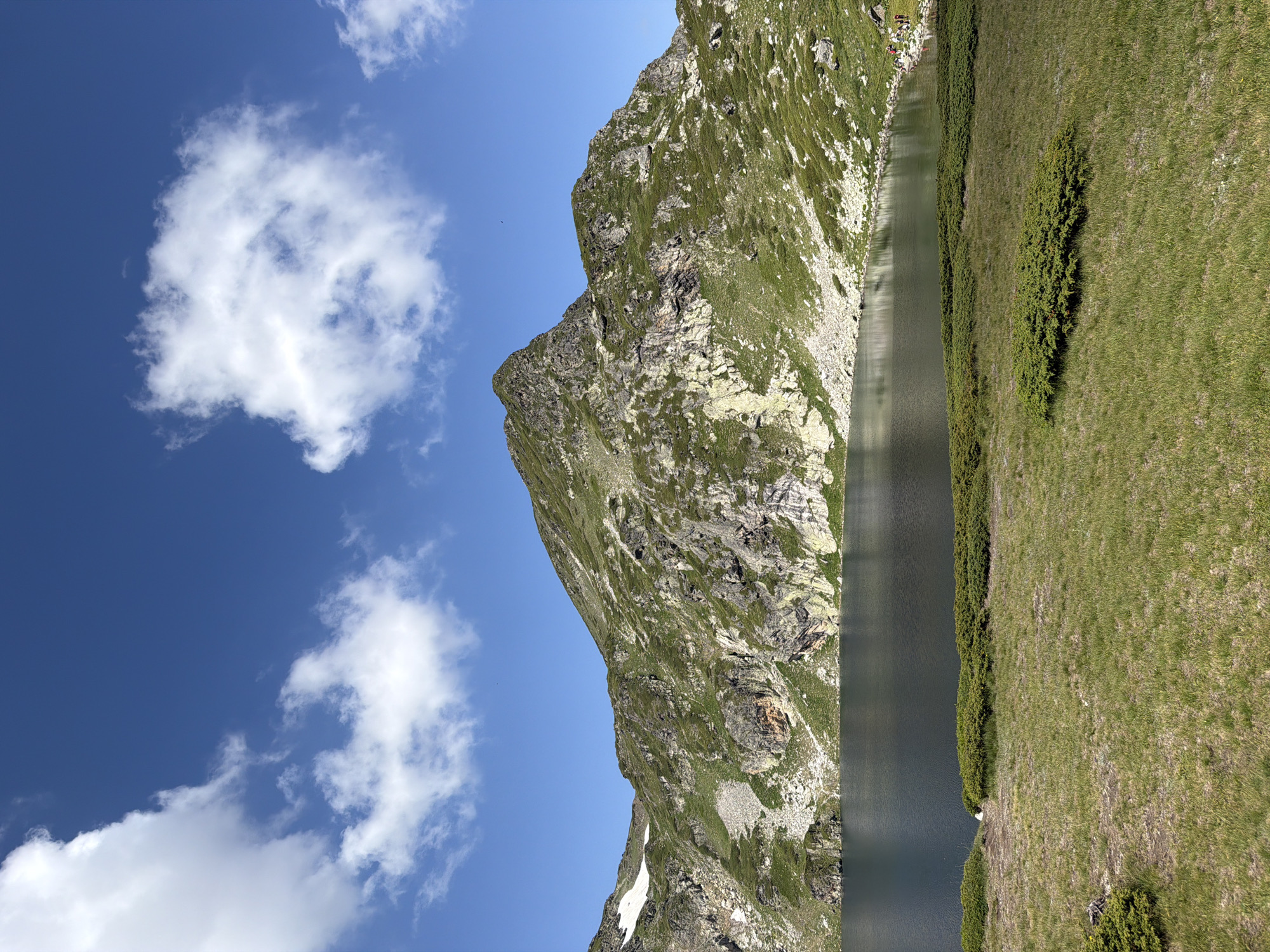The image captures a serene mountain lake scene in Panichishte, Bulgaria, under a bright blue sky dotted with scattered white clouds. The foreground consists of a grassy slope with low-lying green shrubs. Dominating the mid-ground is a dark, still lake reflecting the surrounding landscape. Beyond the lake, a rugged, green mountain rises steeply, its rocky face partially covered in vegetation. A patch of snow is visible on the higher slopes to the left. In the far right of the image, along the shore of the lake, a small group of people, appearing to be tourists, are gathered. The overall impression is one of natural beauty and tranquility.