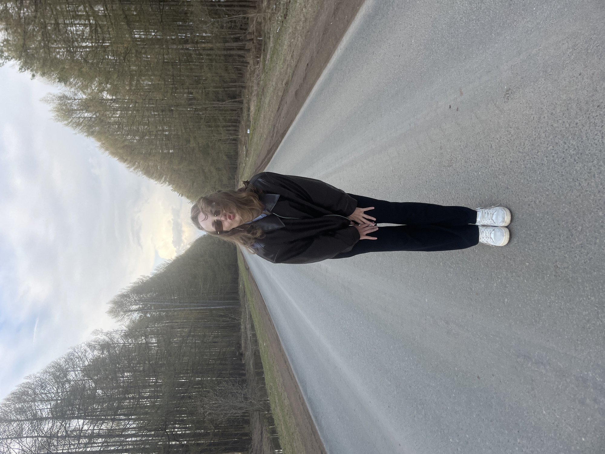 A young woman stands centrally on a paved, two-lane road that stretches into the distance. She is wearing a dark brown or black leather jacket, black pants, and white sneakers, with dark sunglasses obscuring her eyes. Her long, light brown hair falls around her shoulders, and her hands are clasped in front of her. She is positioned facing slightly towards the camera, appearing to pose for a photograph.
The road is made of grey asphalt and is flanked on both sides by dense rows of tall, thin trees that appear to be deciduous and without full foliage, suggesting early spring or late autumn. The trees create a tunnel effect along the road. The sky above is overcast with visible clouds, providing diffused daylight without harsh shadows, possibly indicating an overcast day or late afternoon. The setting is rural or semi-rural. There is no visible text in the image. The location context is Podshivalovo, Russia.